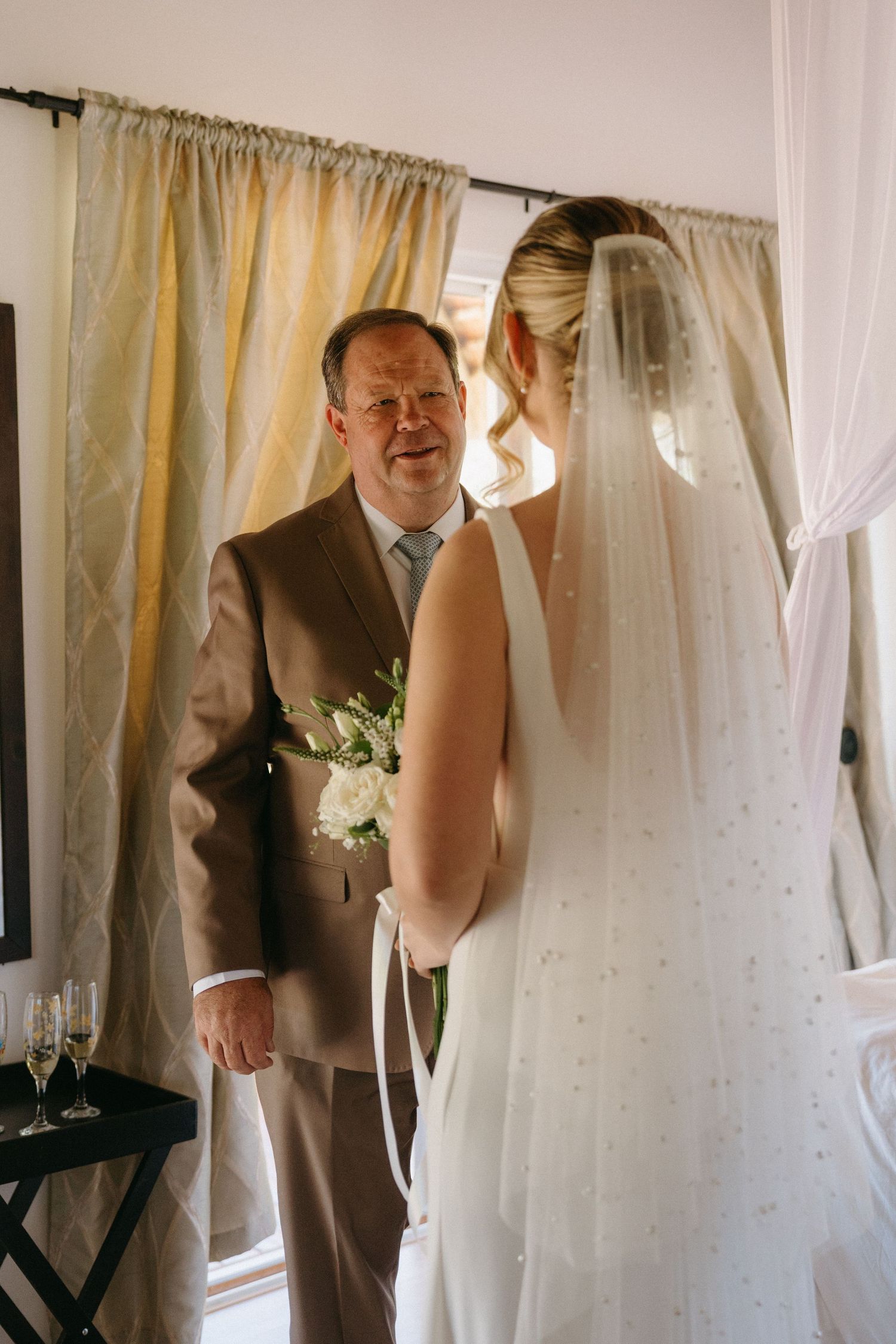 A tender moment between a bride in a flowing white veil and man in a brown suit standing by curtained windows.