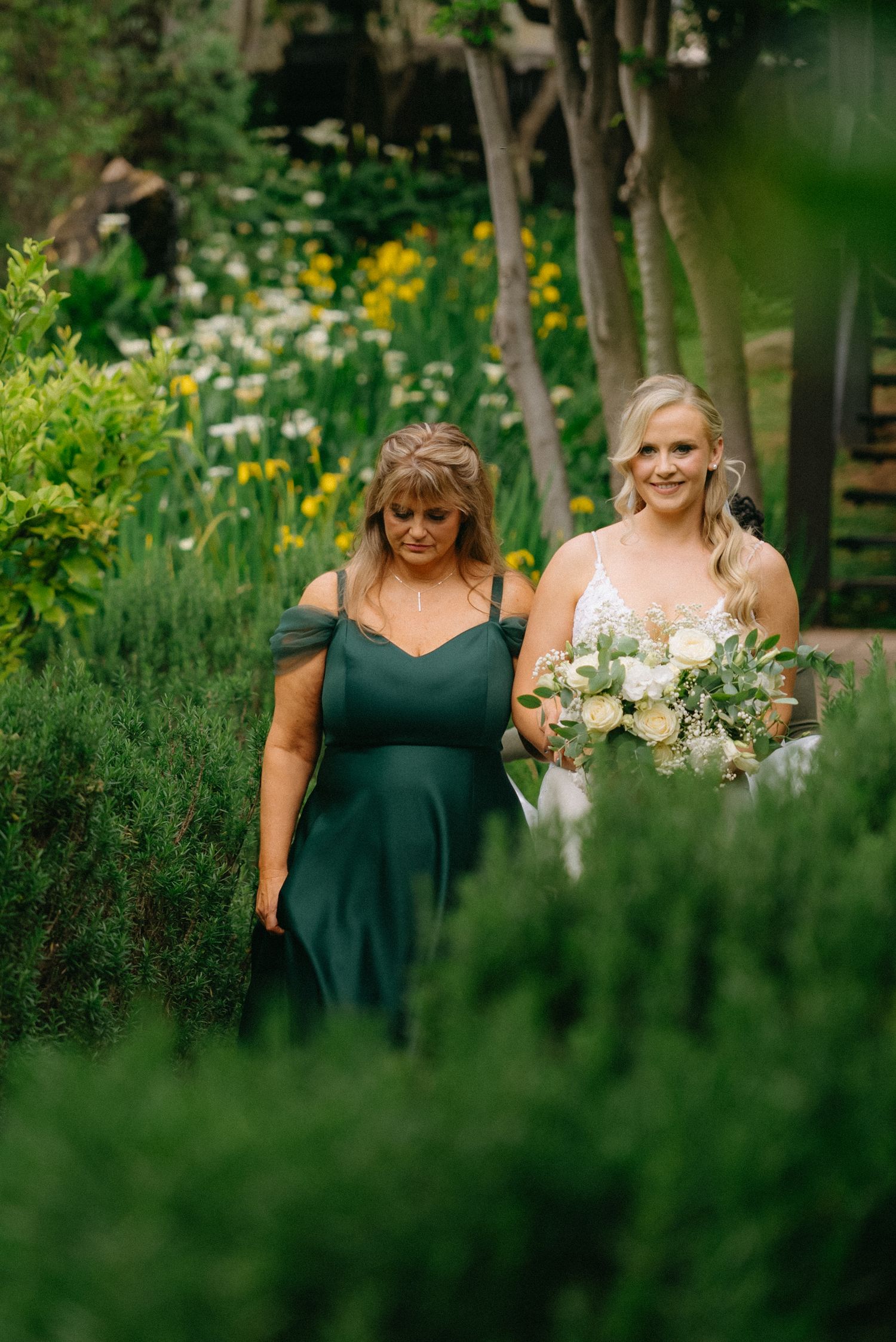 Bridesmaids in emerald green dresses carry white bouquets while walking through a lush garden setting.