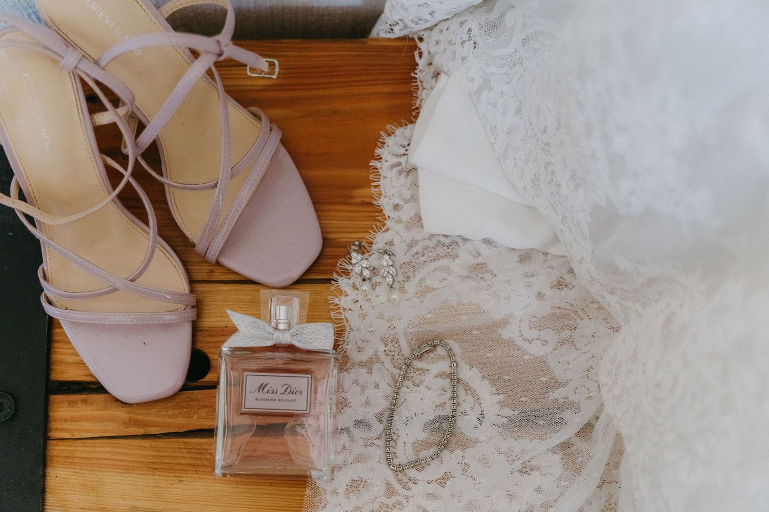 Wedding accessories including blush pink sandals, perfume bottle and delicate lace details on wooden flooring.