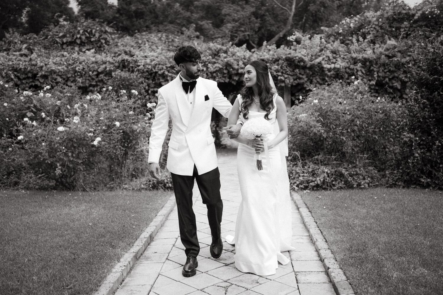 A joyful couple in wedding attire walk together and celebrate on a garden path in black and white.