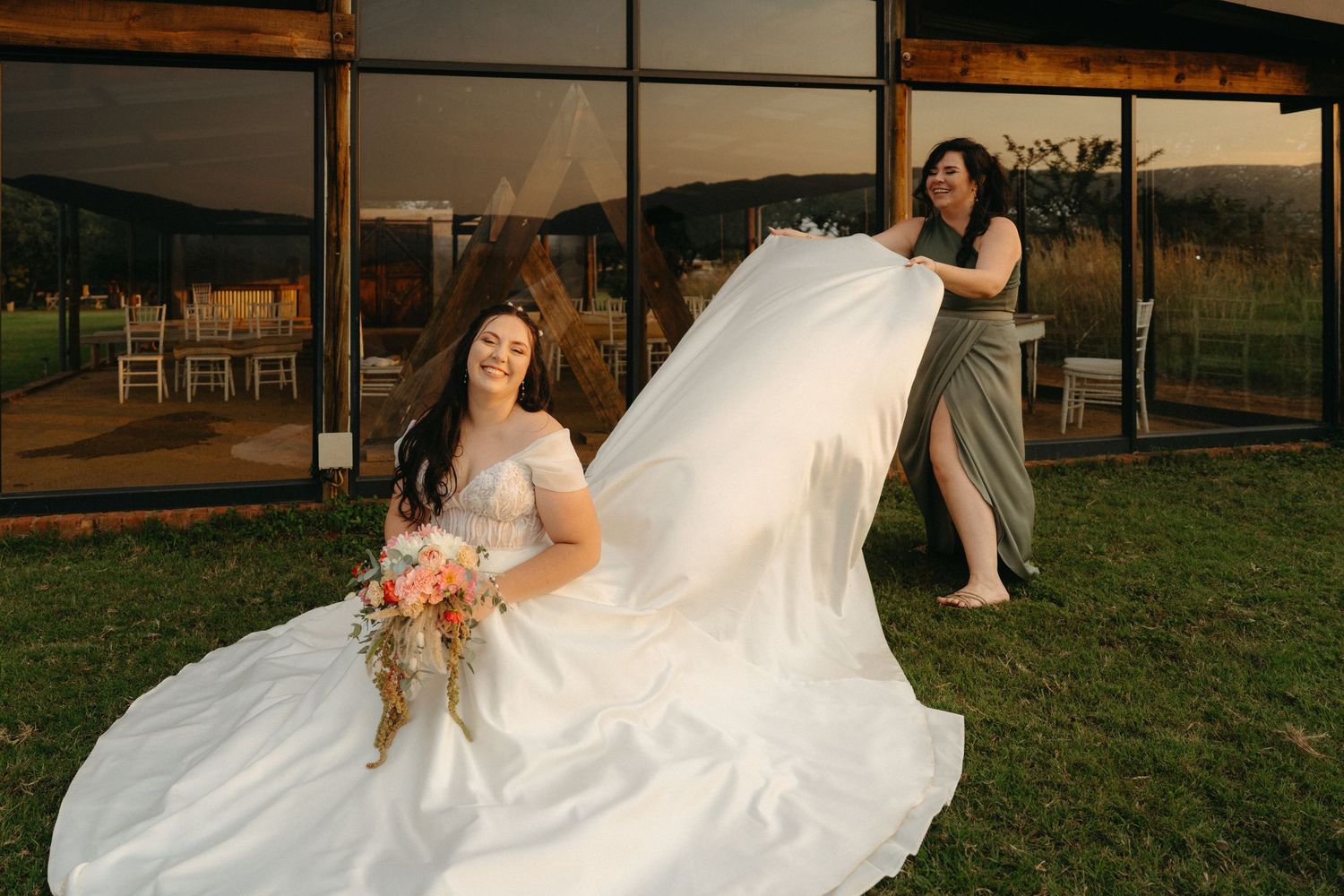 A bride in a flowing white wedding dress sits while her veil billows in the wind during sunset.