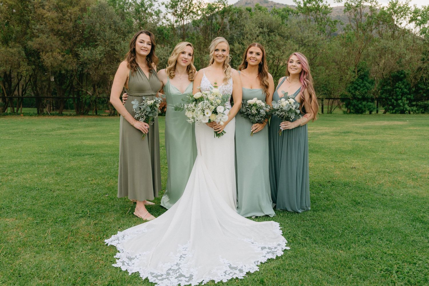 Wedding party in gray bridesmaid dresses and white bridal gown pose together on a grassy lawn.