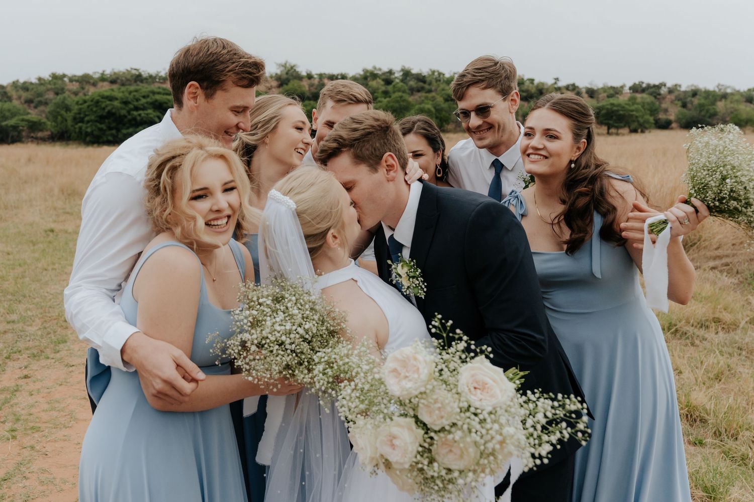 Wedding party laughs together while holding white floral bouquets in an outdoor field setting.