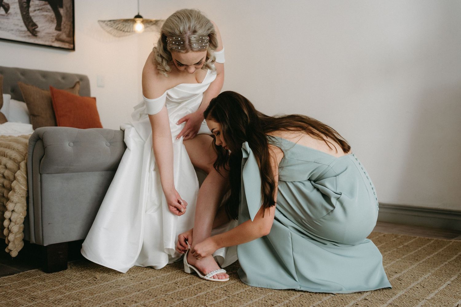 A bridesmaid in blue helps adjust the bride's shoes during wedding preparation.