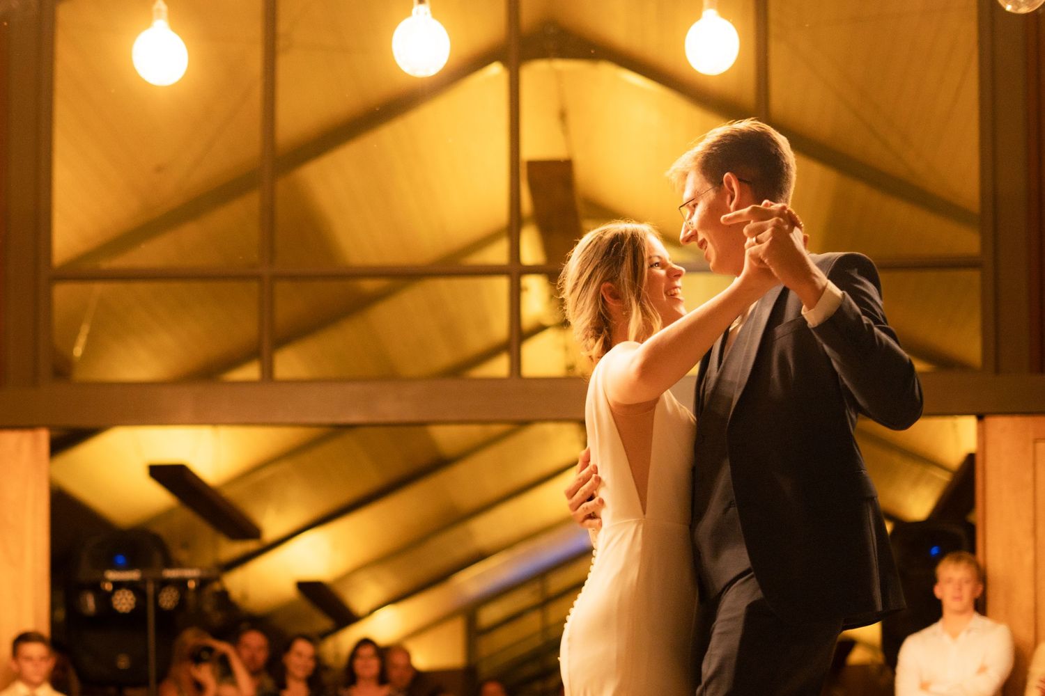 A couple shares their first dance at their wedding reception under warm glowing lights in a rustic venue.
