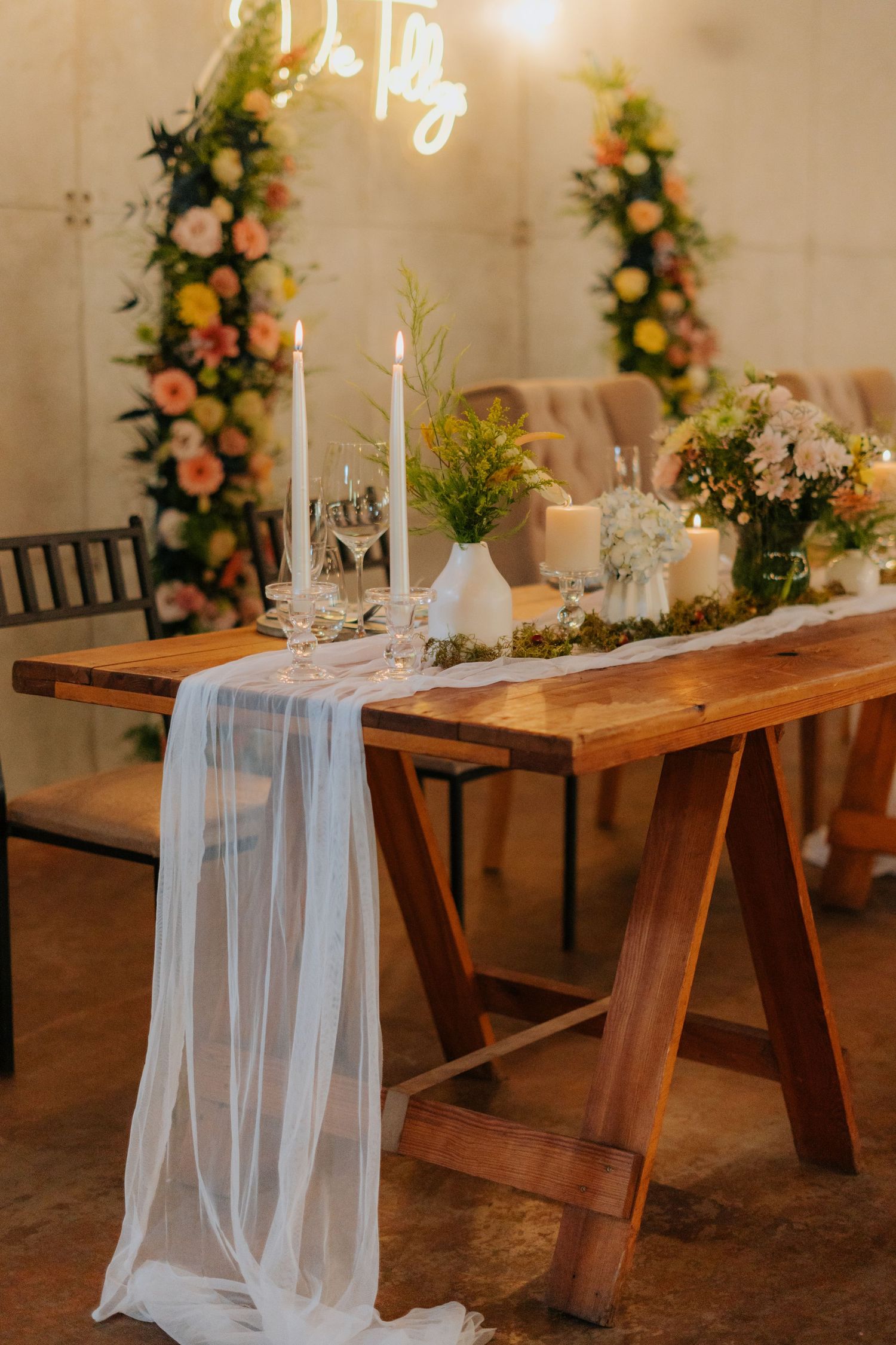 Rustic wooden table decorated with white tulle runner, candles and floral arrangements for an elegant wedding reception setup.