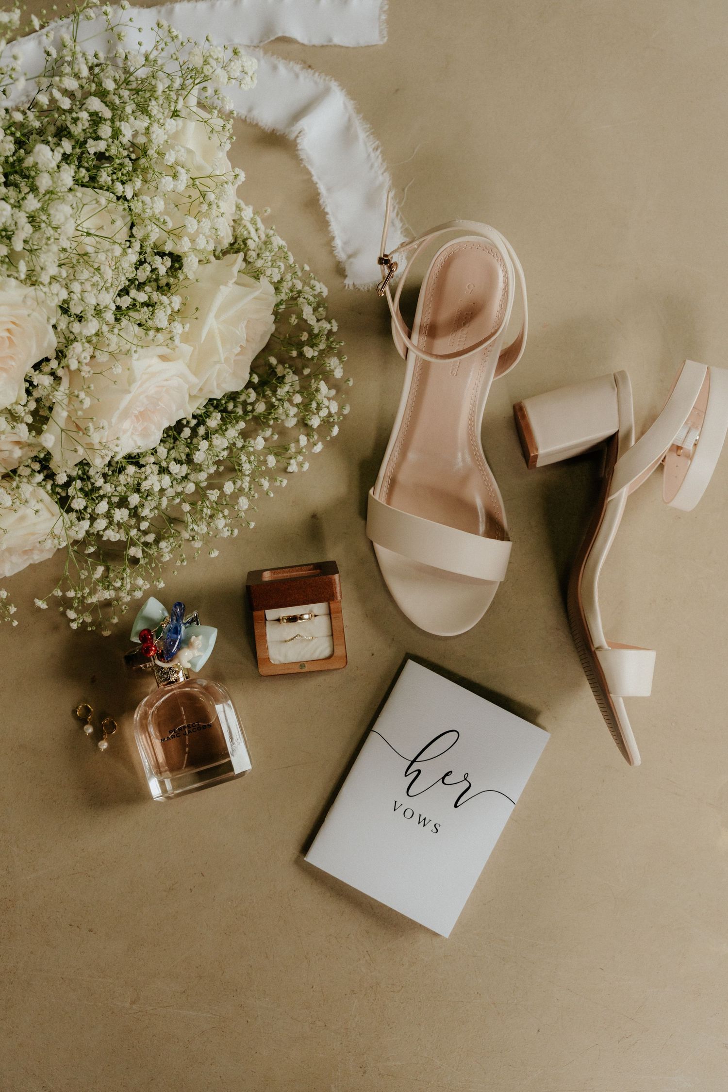 Wedding details flatlay with white flowers, perfume bottle, pale sandals and vow book on beige background.