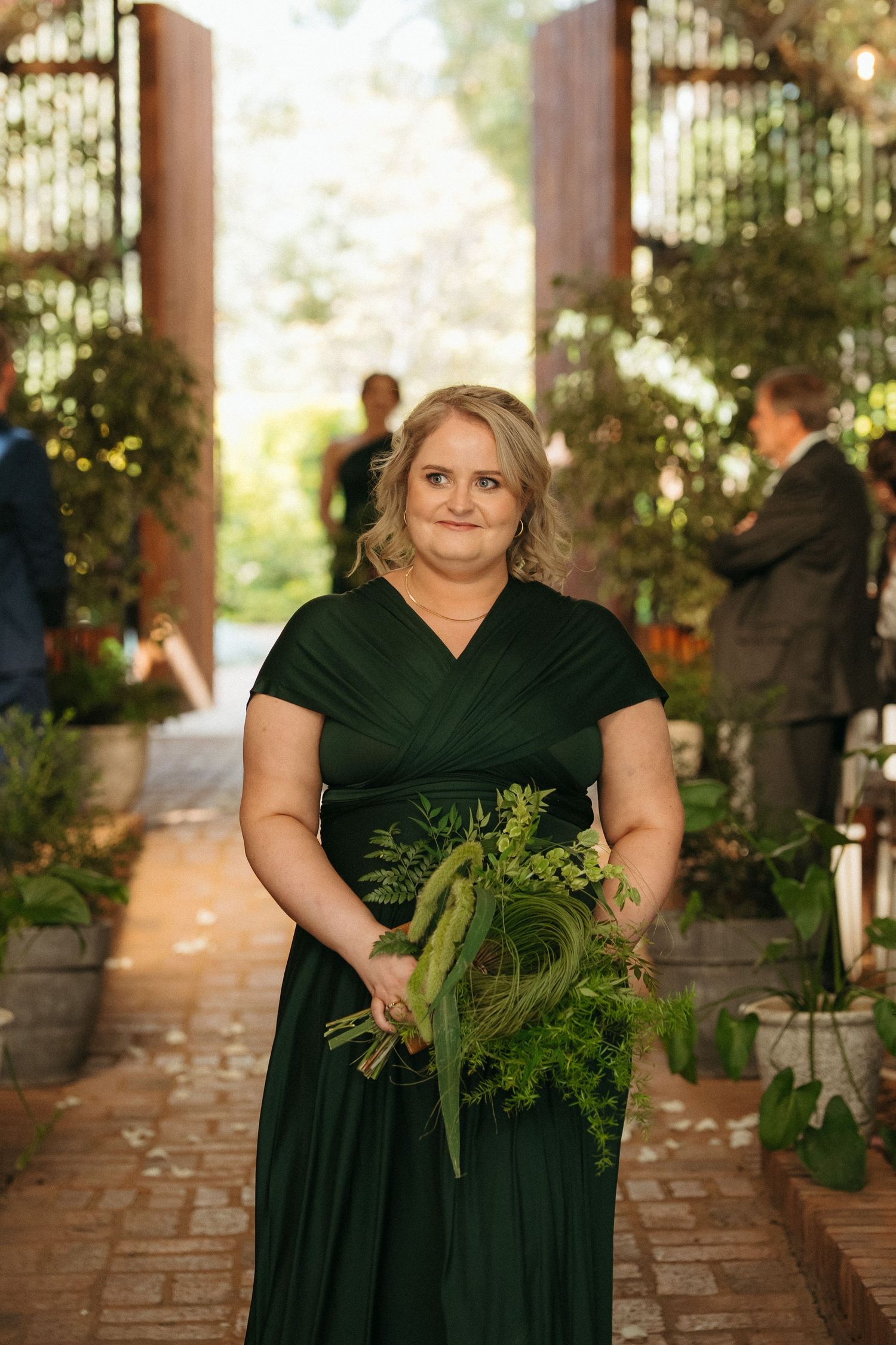 Bridesmaid in dark green wrap dress holding lush greenery bouquet stands in garden wedding venue aisle.