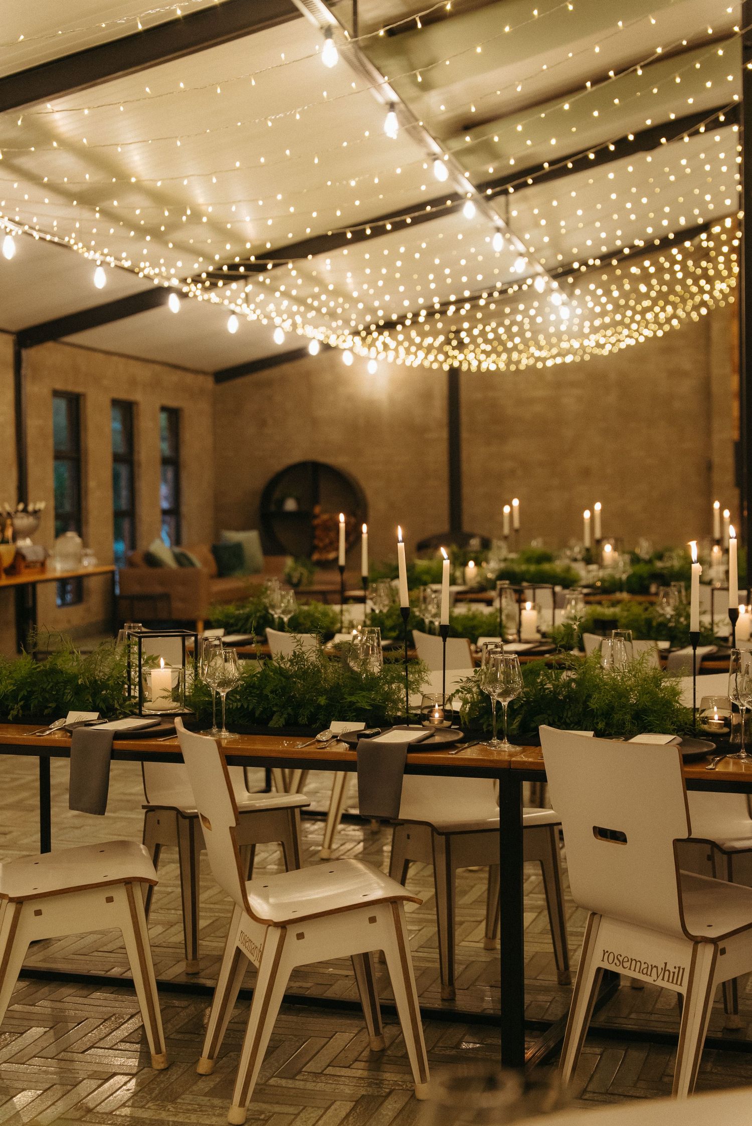 Elegant wedding reception with string lights overhead, white chairs, candles, and lush greenery centerpieces on wooden tables.