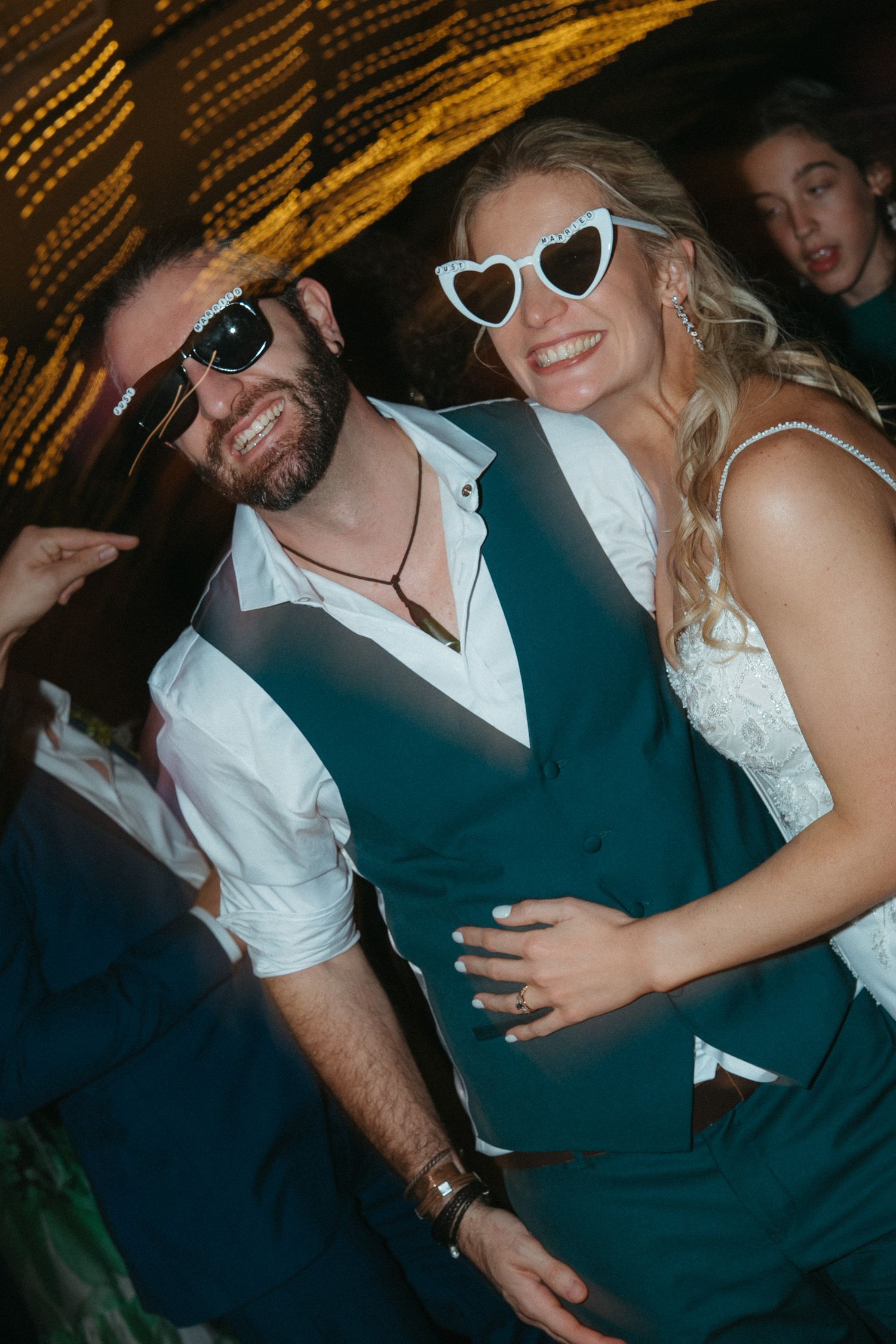 A smiling couple in wedding attire wearing fun sunglasses pose together at a wedding reception party.