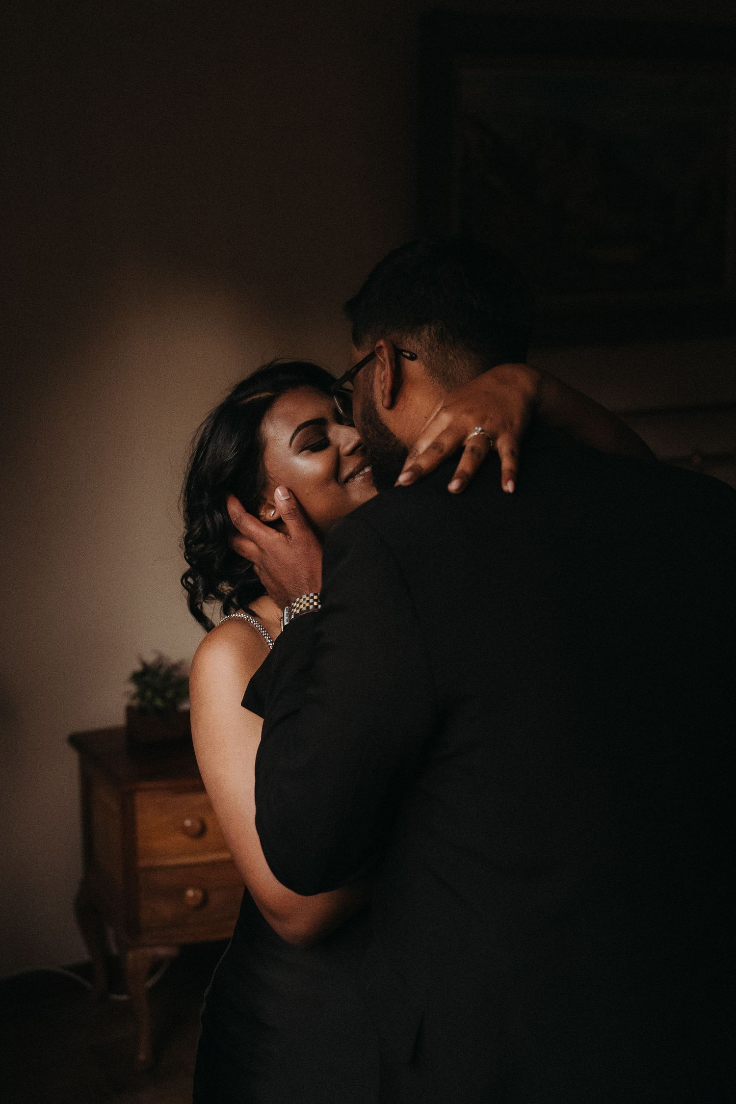 A couple shares an intimate embrace in a dimly lit room, the woman smiling softly while wearing a pearl bracelet.