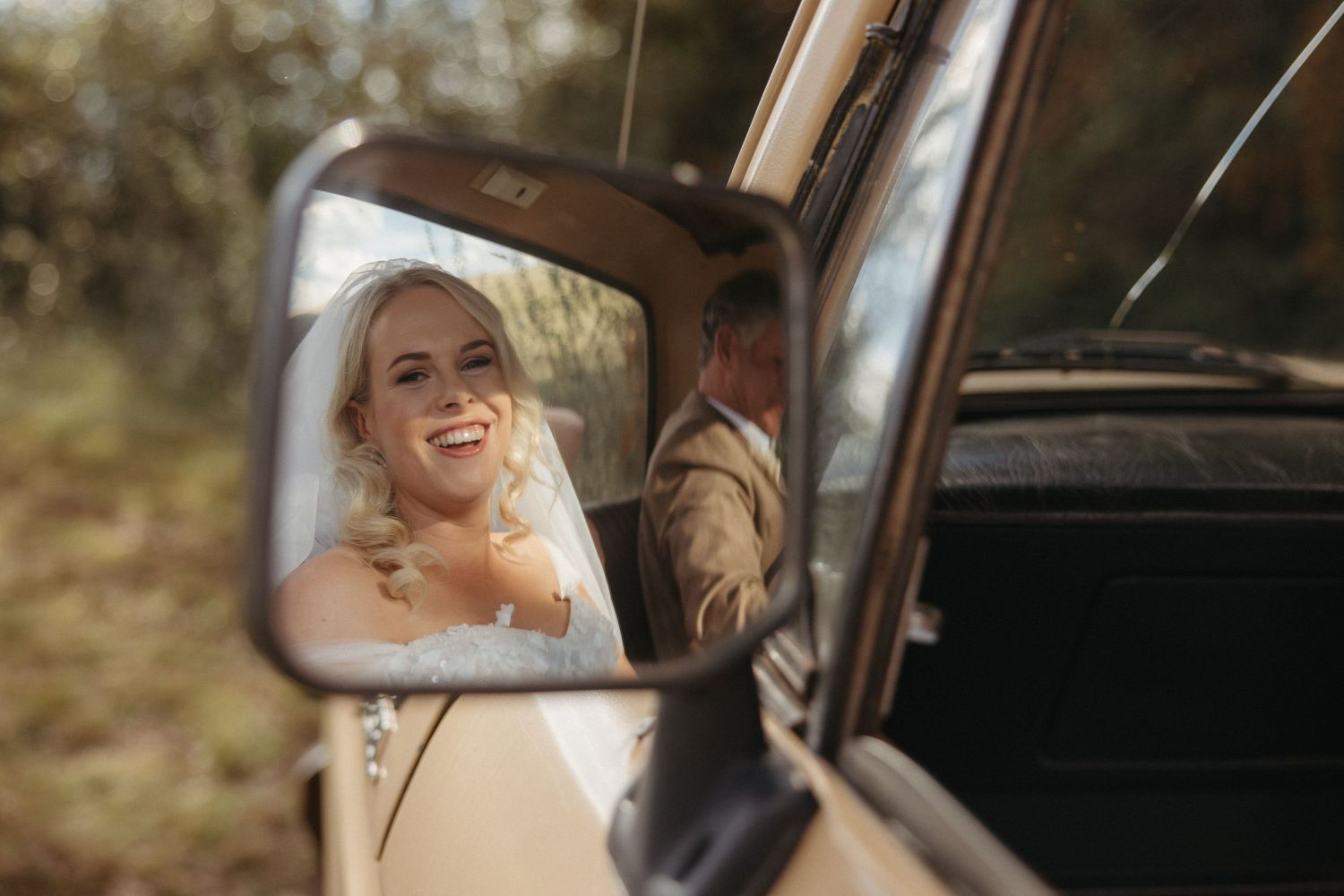 Bride smiling joyfully reflected in vintage car side mirror on her wedding day.