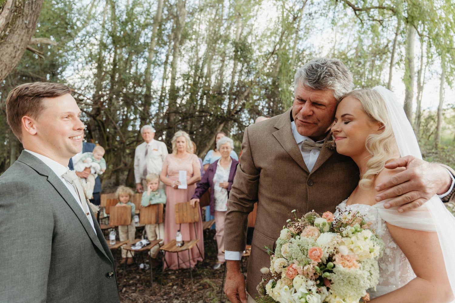 Bride in veil with bouquet hugged by father in tan suit, groom watching, guests gathered outdoors at woodland wedding.