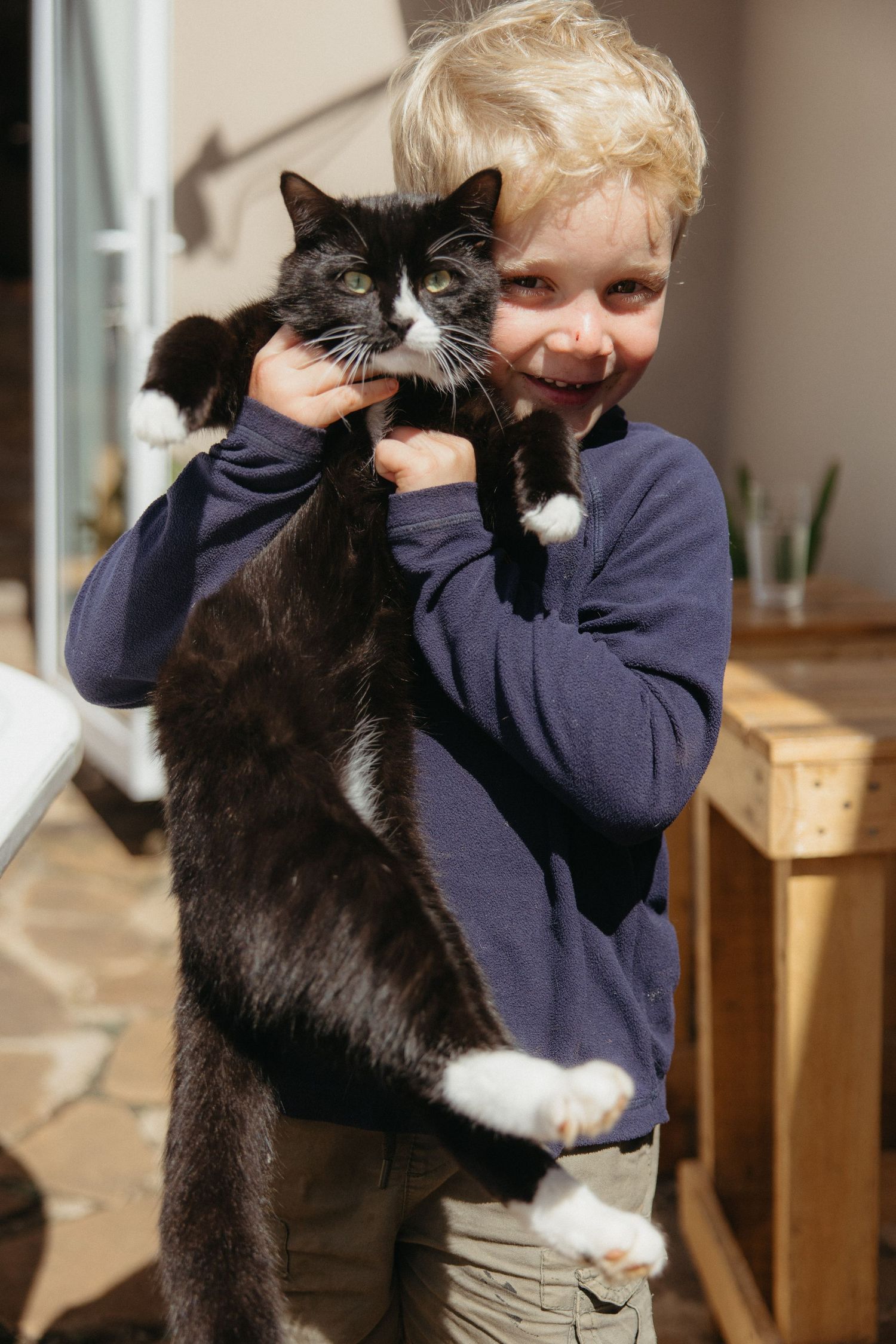 Smiling blonde boy holding a large black and white tuxedo cat outdoors in sunny patio setting.