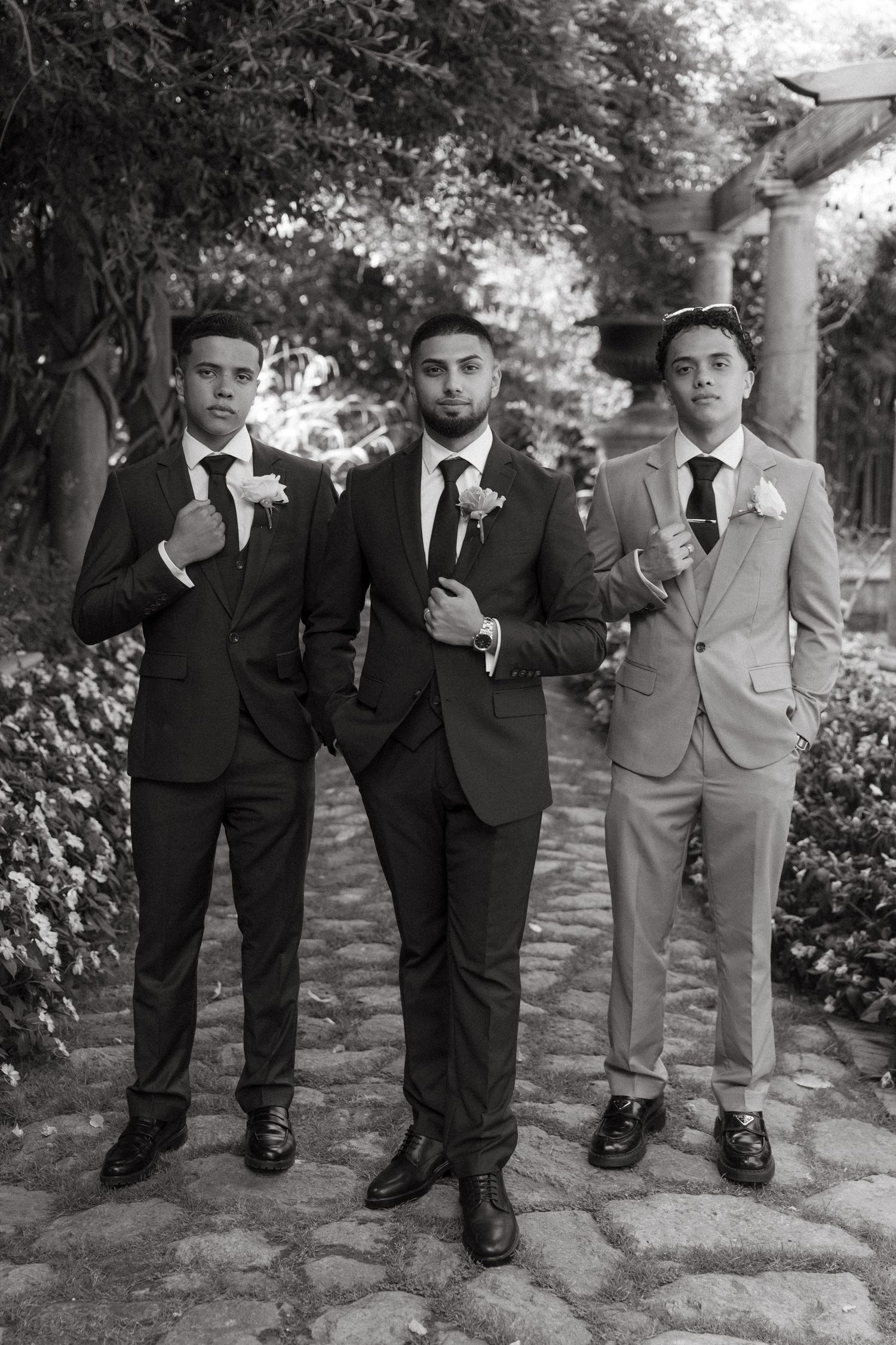 Three groomsmen in suits with boutonnieres pose on a stone path surrounded by lush greenery in a black and white photo.