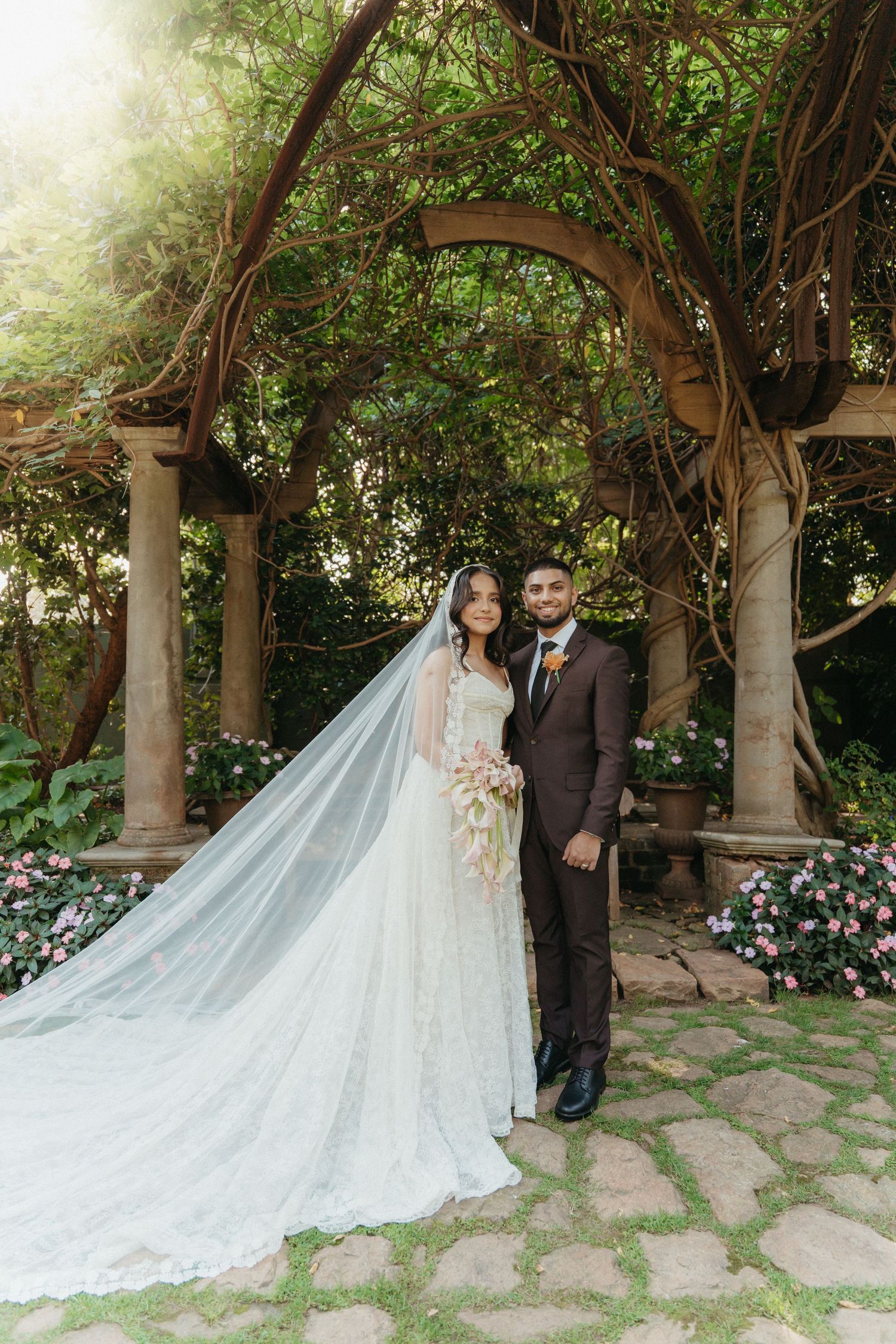 Bride and groom pose under vine-covered stone archway in Beechwood Gardens, bride wearing cathedral veil and lace gown.