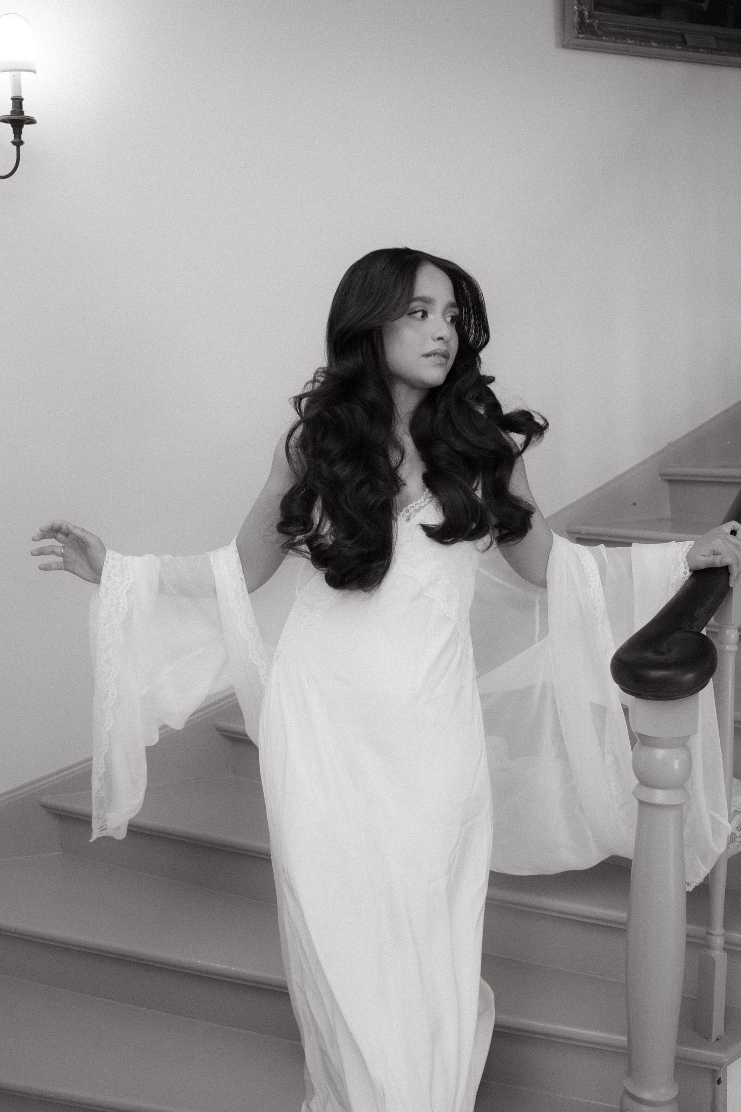 Black and white photo of a woman in a flowing white gown with bell sleeves on a staircase.