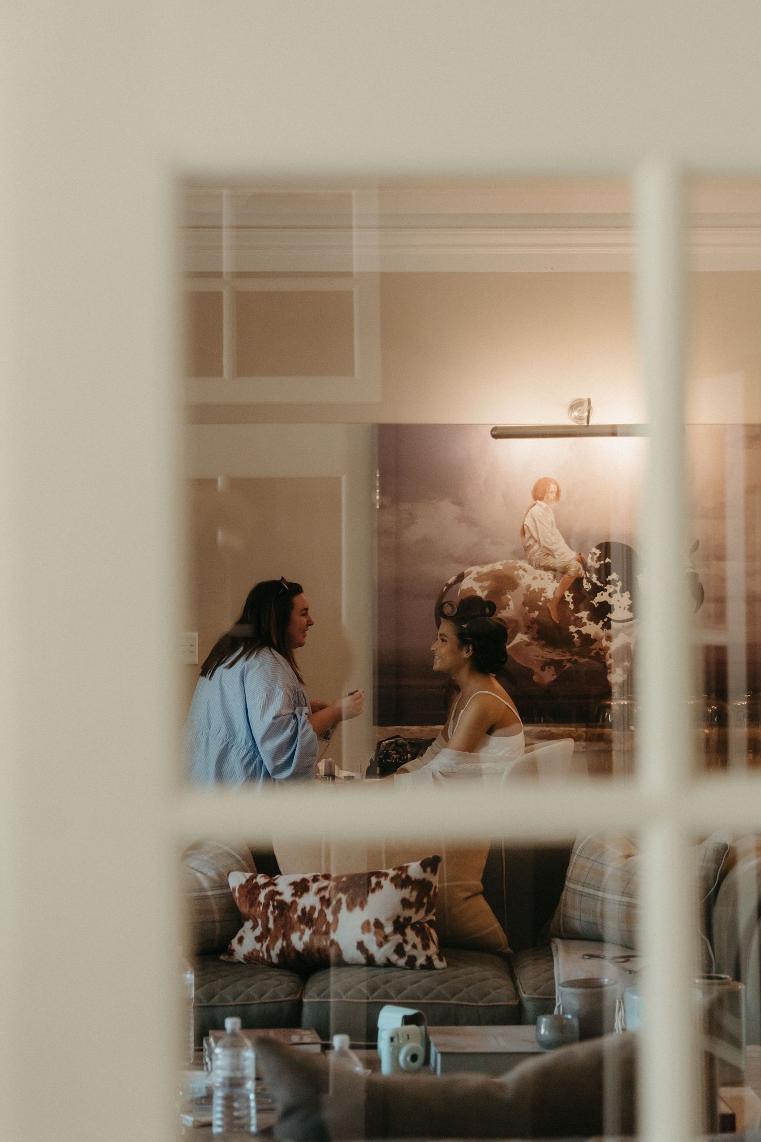Two women conversing in a warmly lit living room with a large artwork on the wall, viewed through a window.