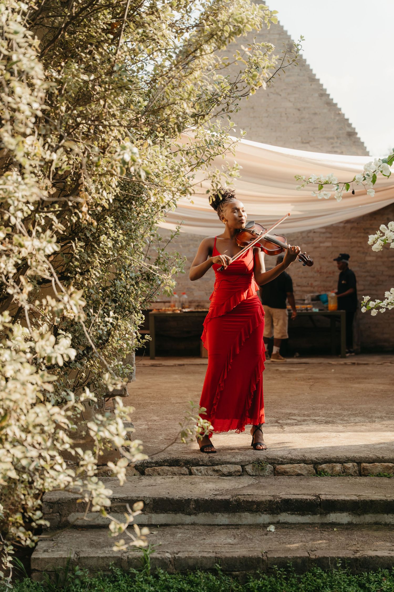 A woman in a red dress plays violin outdoors at an elegant garden event, framed by white flowering branches.