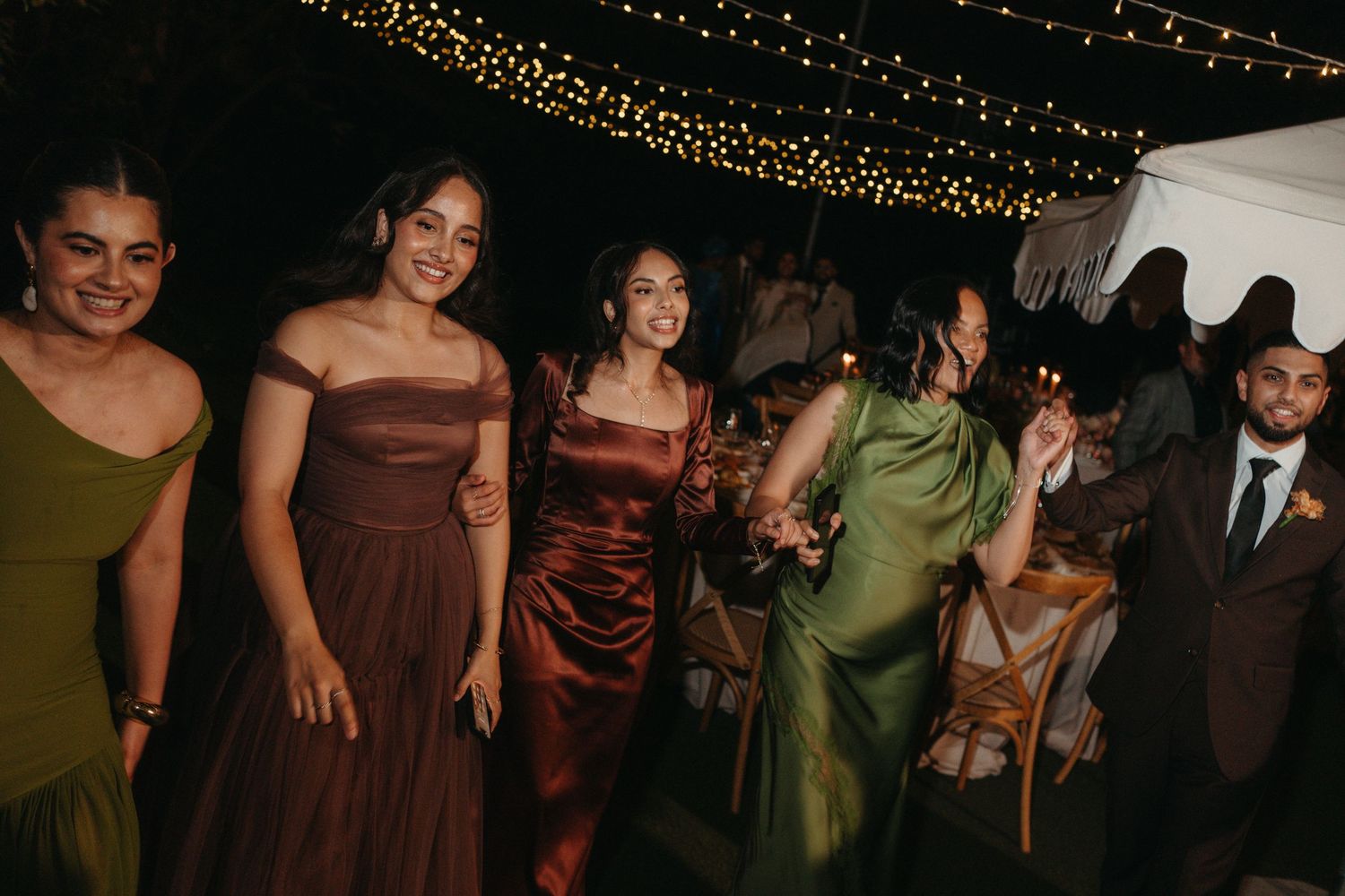 Guests in elegant satin dresses dance joyfully at an outdoor wedding reception under warm string lights at night.