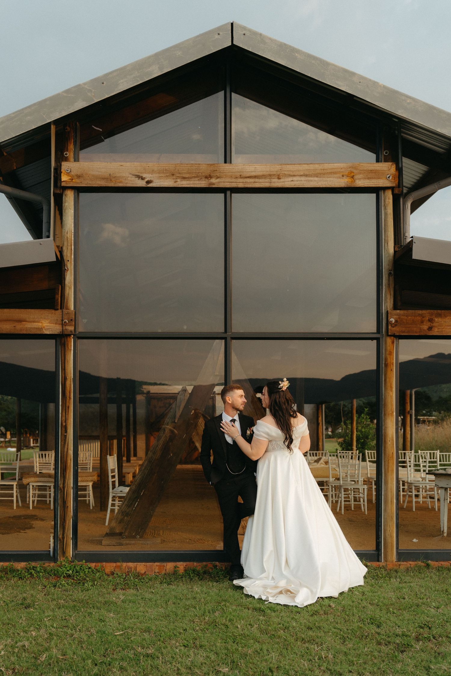 Modern glass wedding venue with wooden beams showcases couple in elegant attire against mountain scenery.