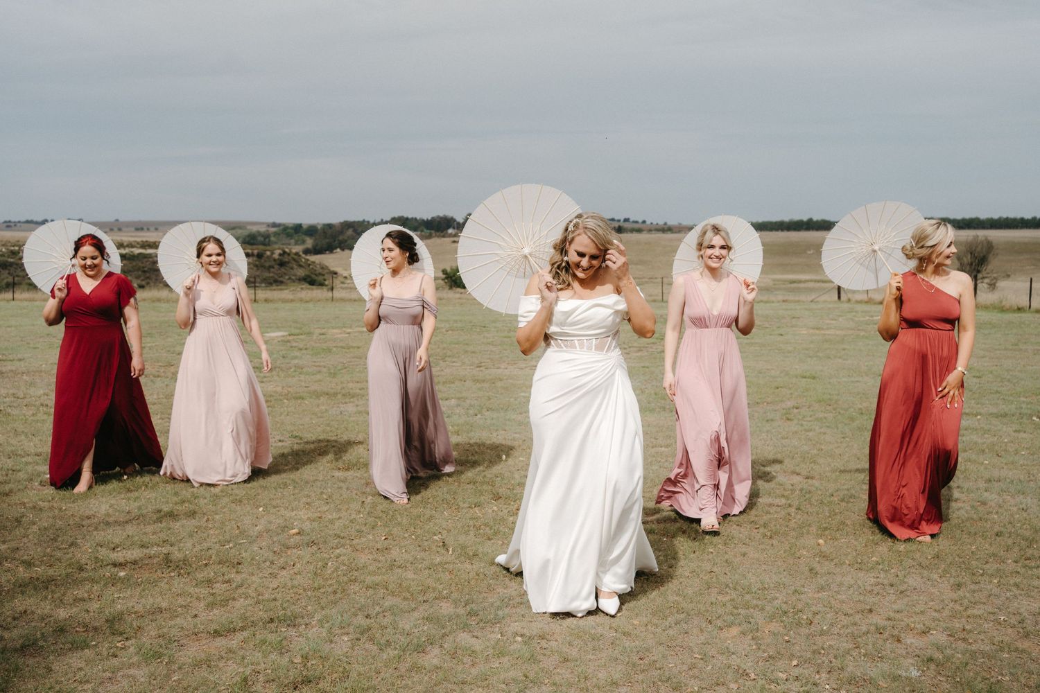 Wedding party posing with white parasols in an outdoor field setting.