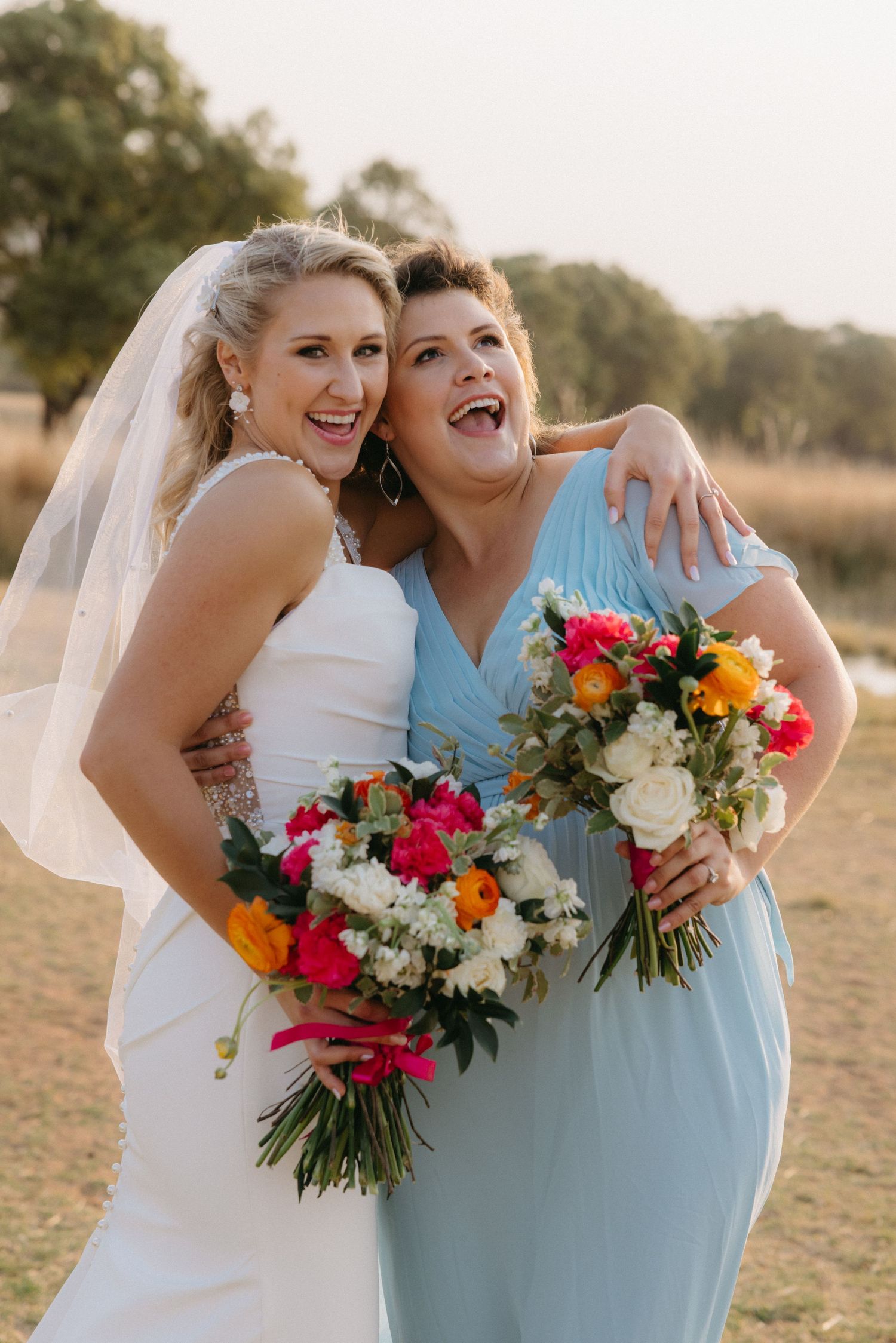Two people in wedding attire holding vibrant floral bouquets in an outdoor setting at sunset.