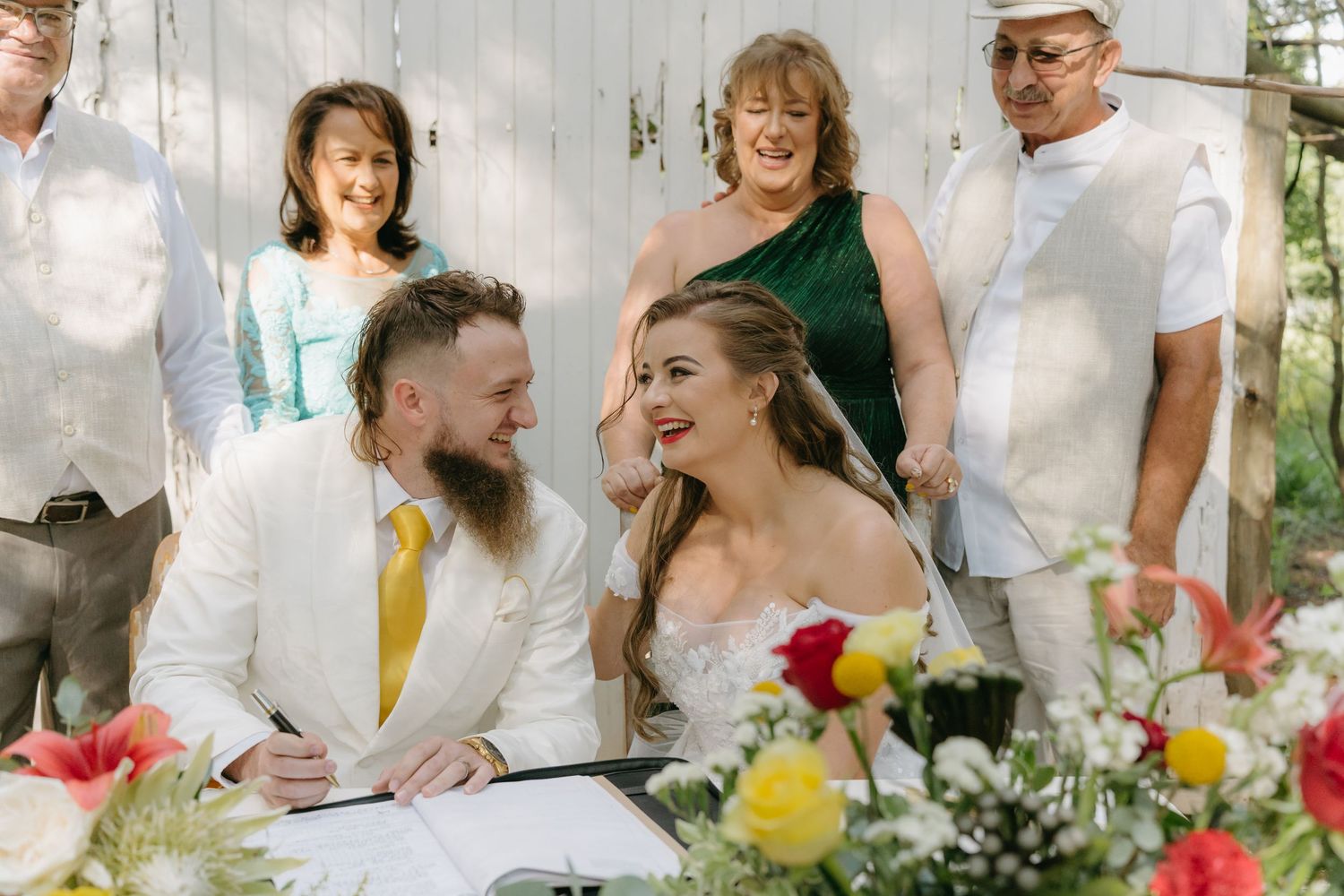 Wedding party gathered at a rustic outdoor reception with cheerful smiles and colorful floral arrangements.