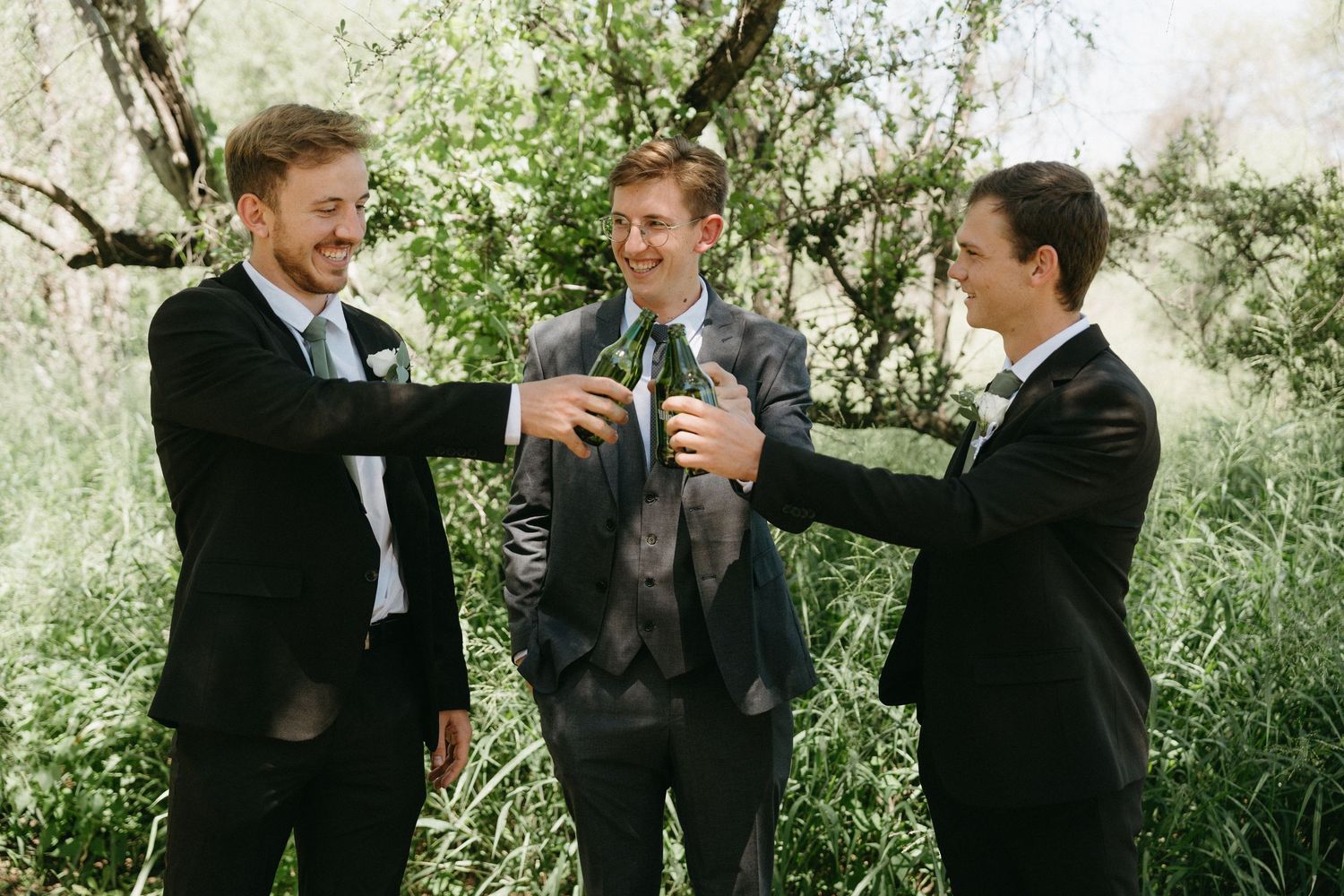 Three groomsmen in suits toast with drinks while celebrating outdoors among trees.