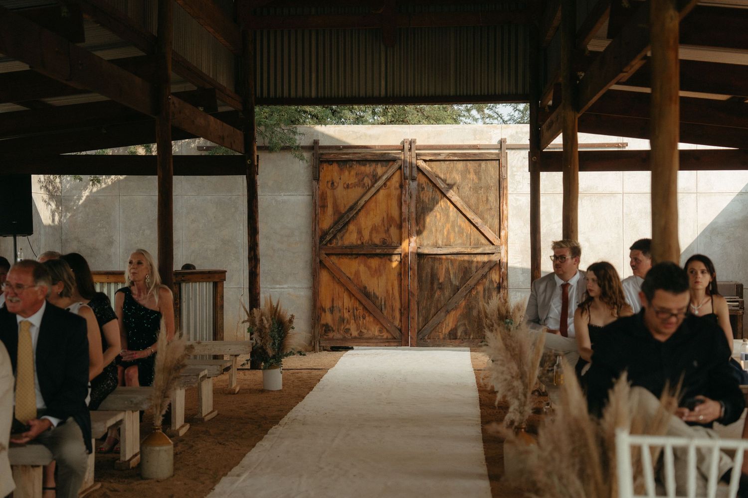 Wedding guests seated in rows inside a rustic barn venue with wooden doors and dried pampas grass decorations.
