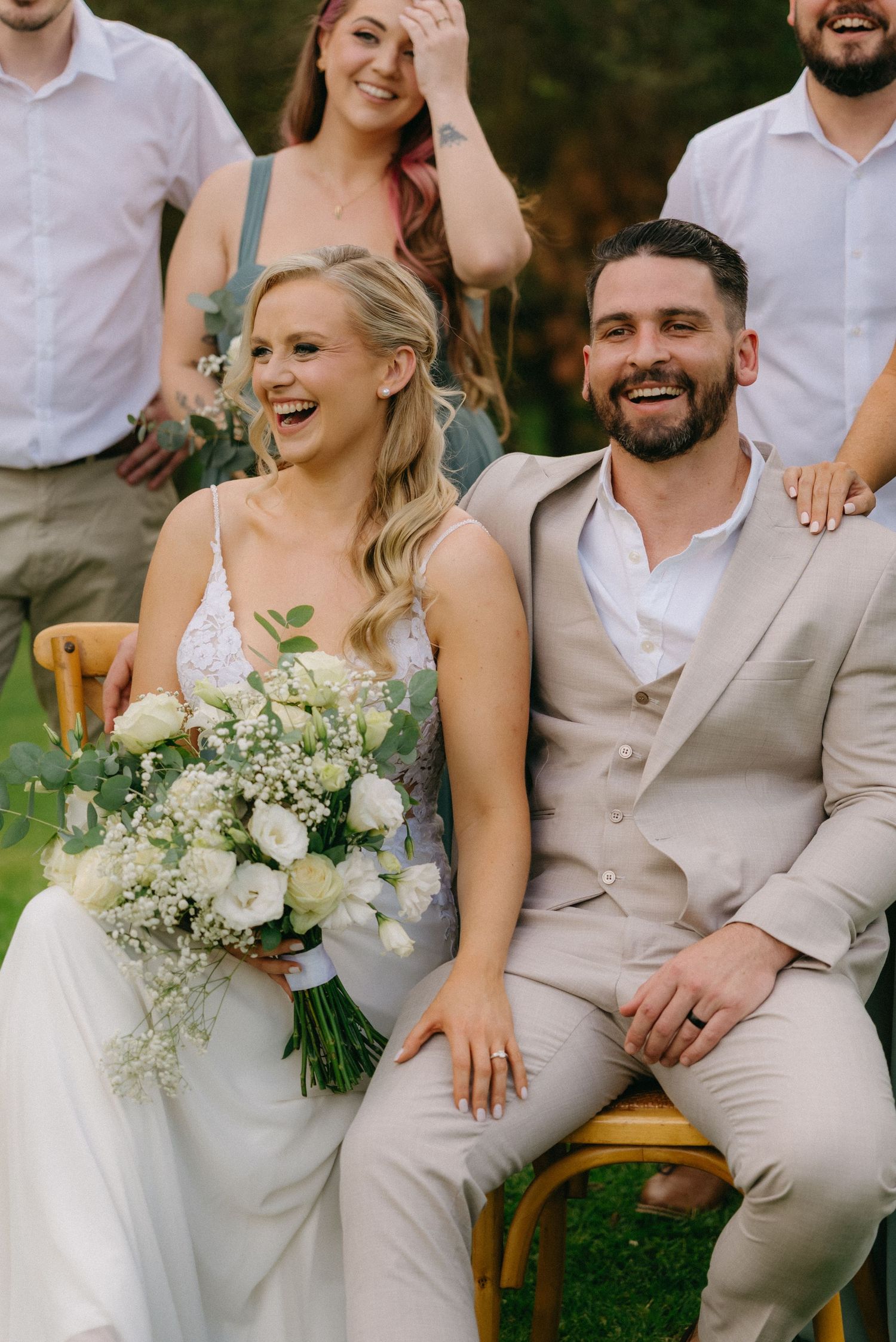 Wedding party seated outdoors sharing joyful moments with white floral bouquet.