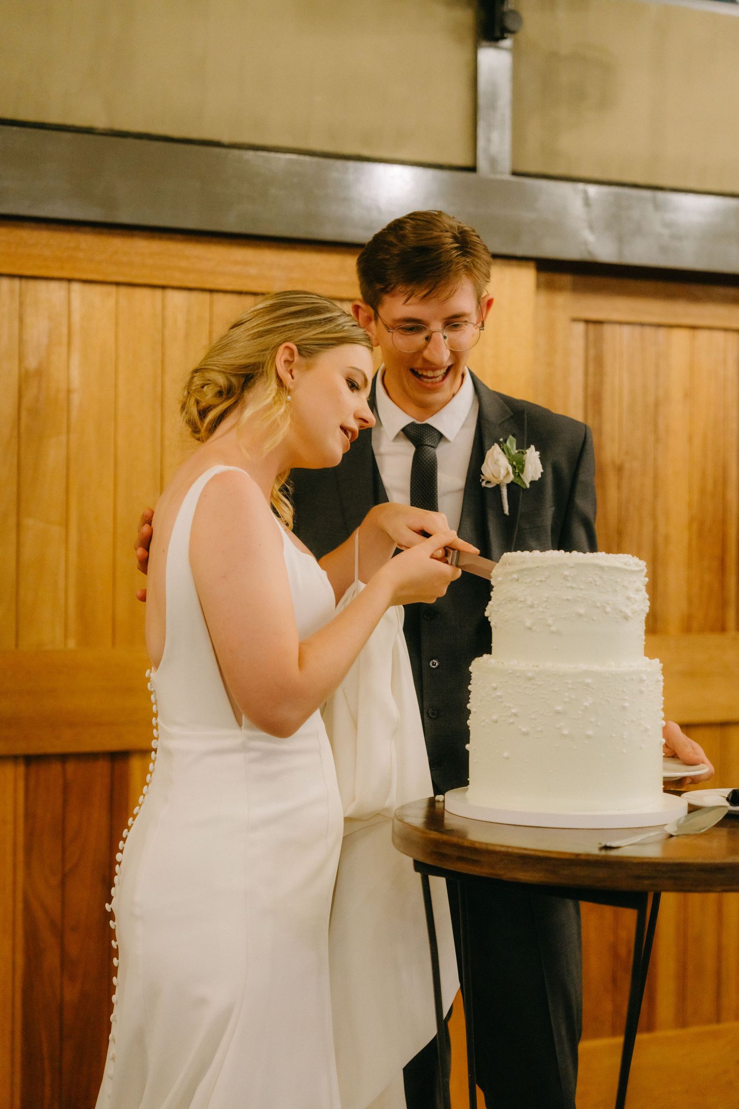 A couple shares a joyful moment while cutting their elegant white two-tiered wedding cake.