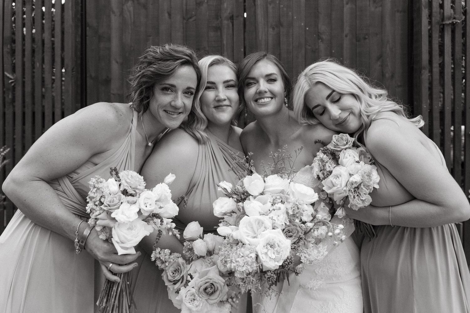Four bridesmaids in matching neutral dresses pose together with white floral bouquets against a rustic wooden wall.