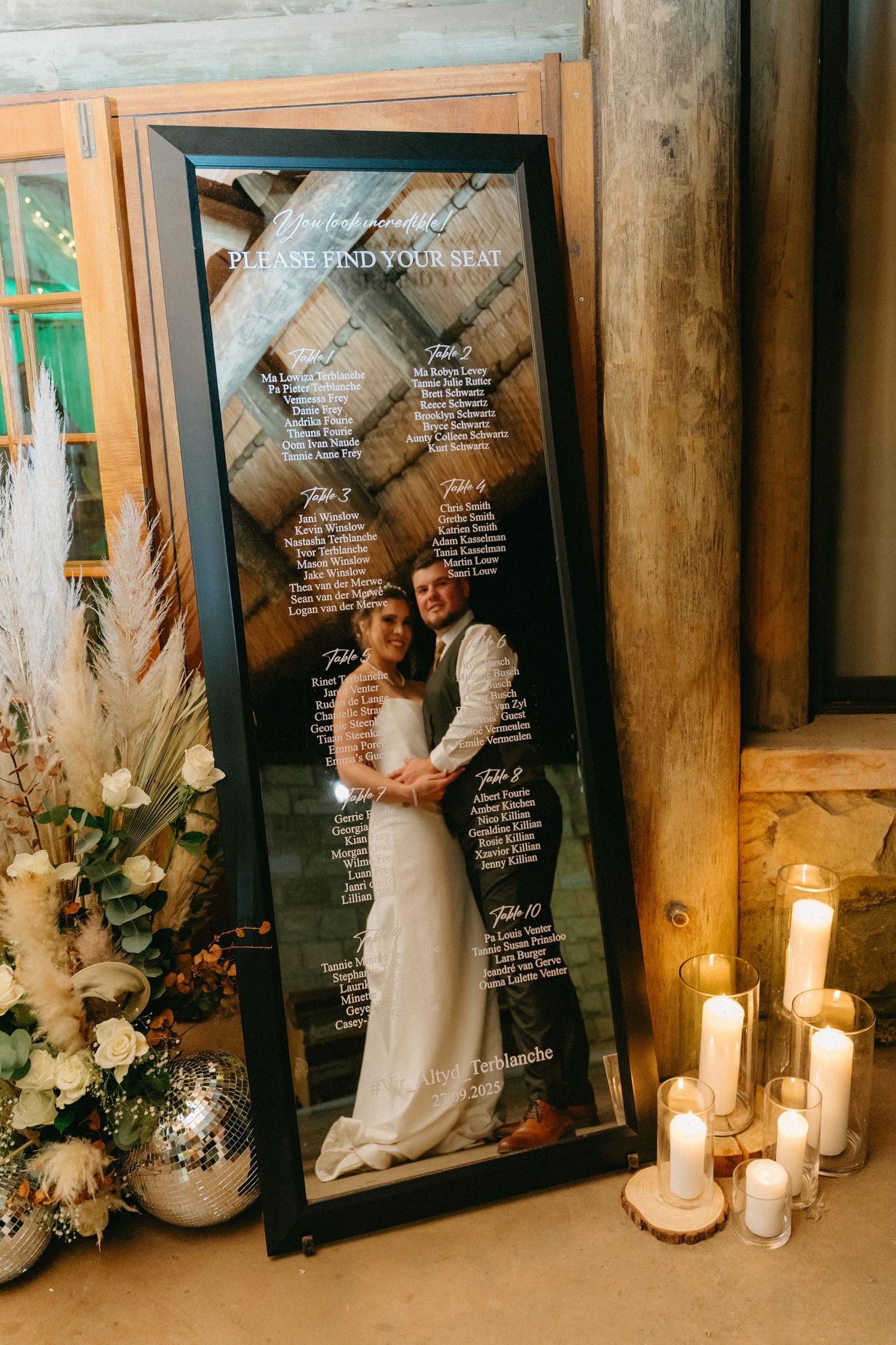 Wedding seating chart display with mirrors, candles and floral arrangements in rustic wooden setting.