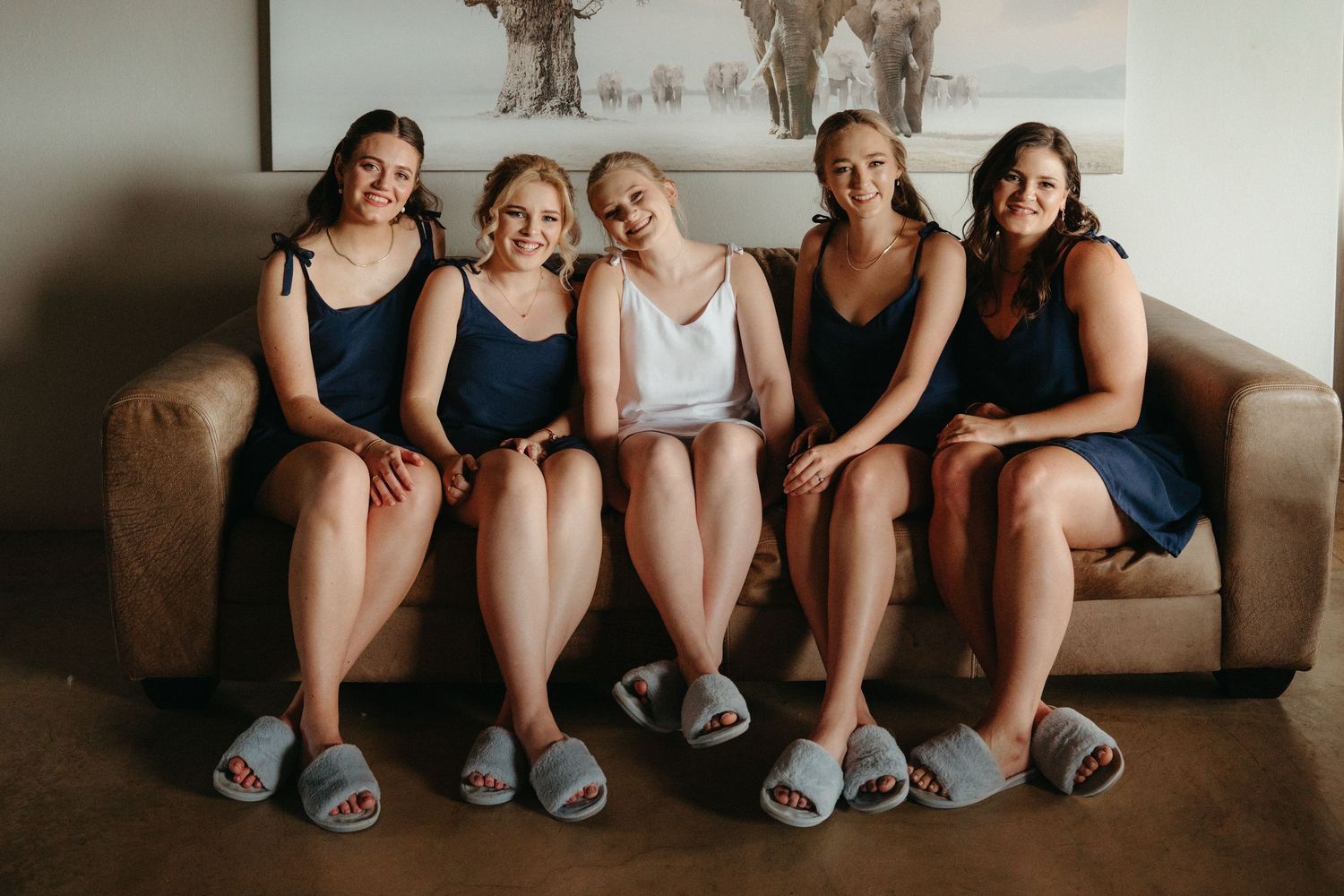 Wedding party members sitting together on a brown couch wearing matching slippers.