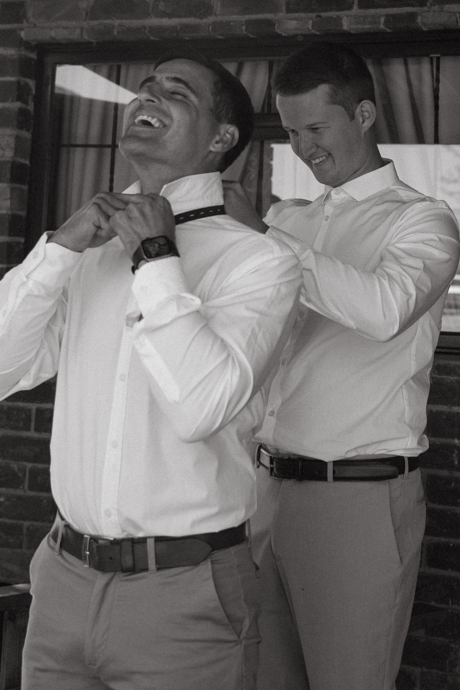 Groomsmen in white dress shirts and gray pants share a lighthearted moment together before the wedding ceremony.