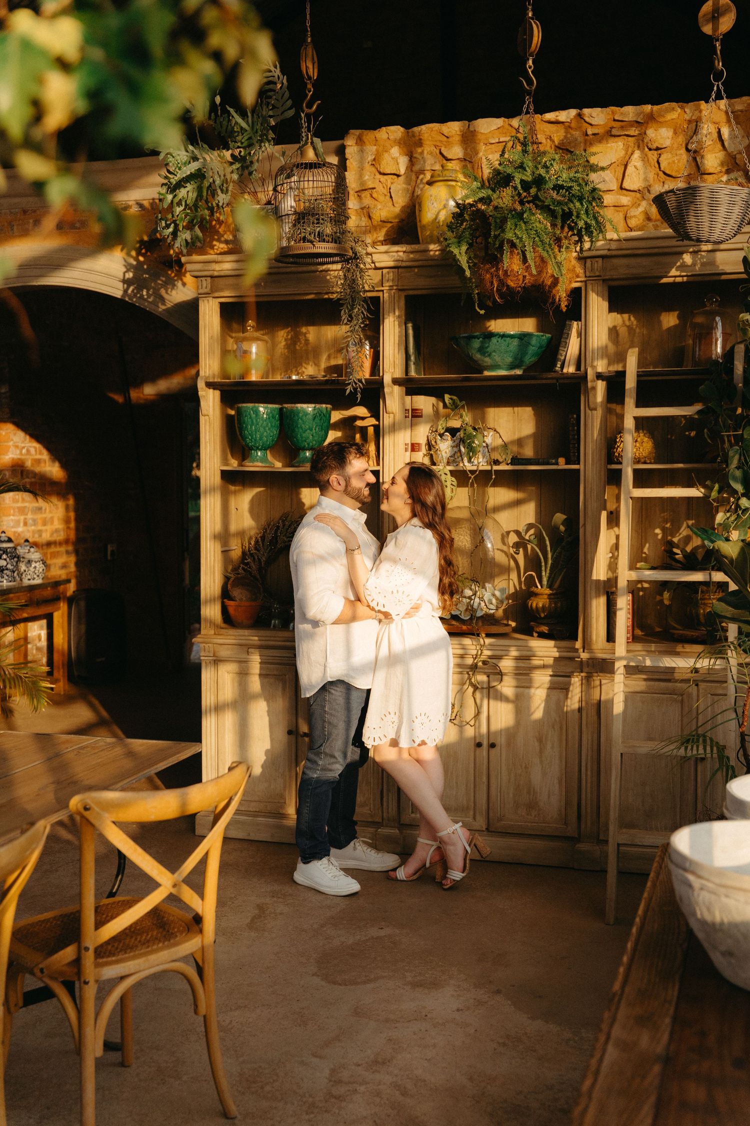 Romantic couple in white clothing sharing an intimate moment among hanging plants in rustic interior.