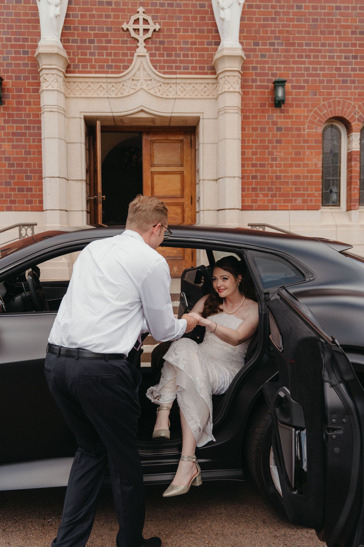 A bride in a white wedding dress is being helped into a black car outside a brick building.