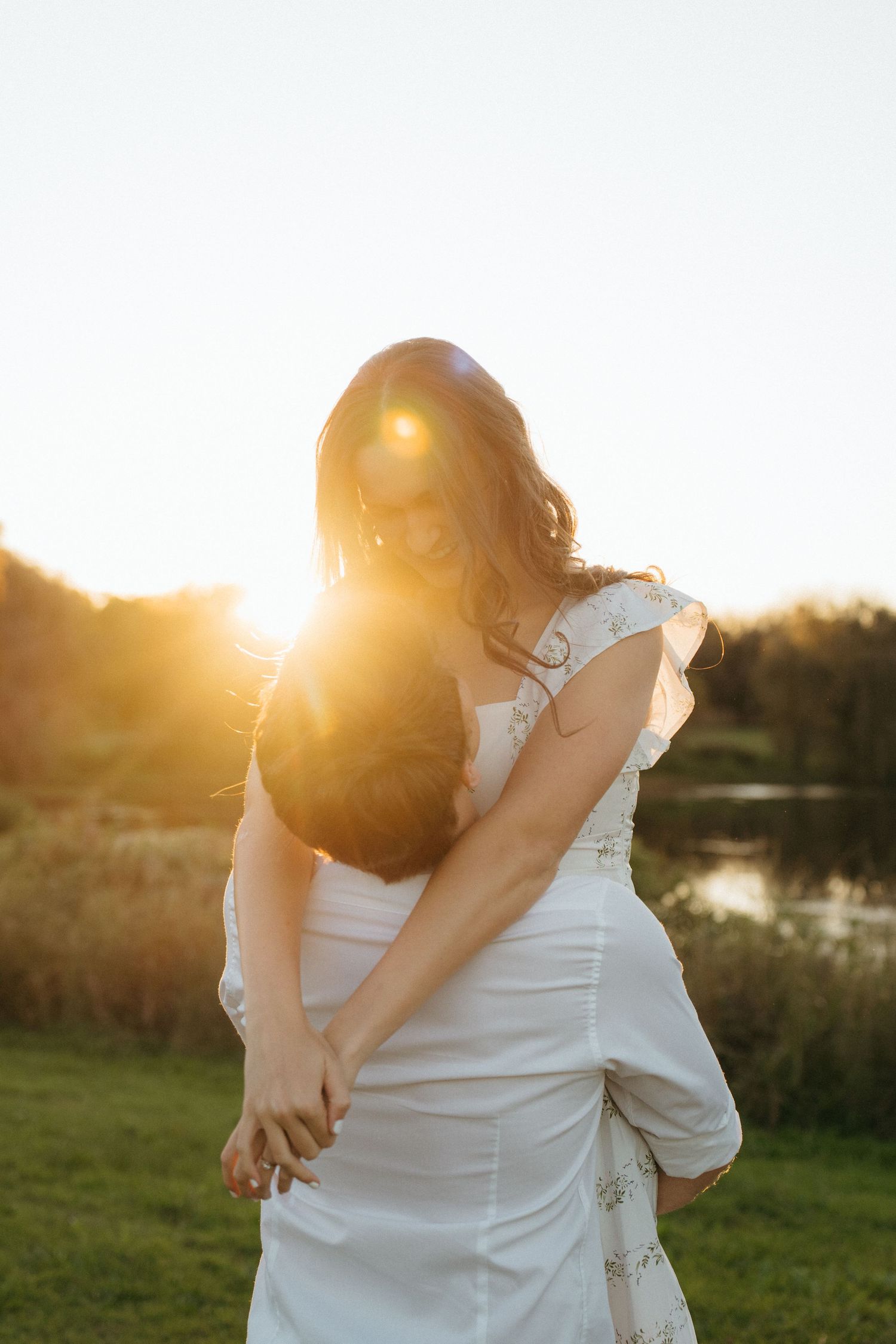 A romantic sunset embrace between a couple wearing white, creating a dreamy backlit silhouette by a lake.