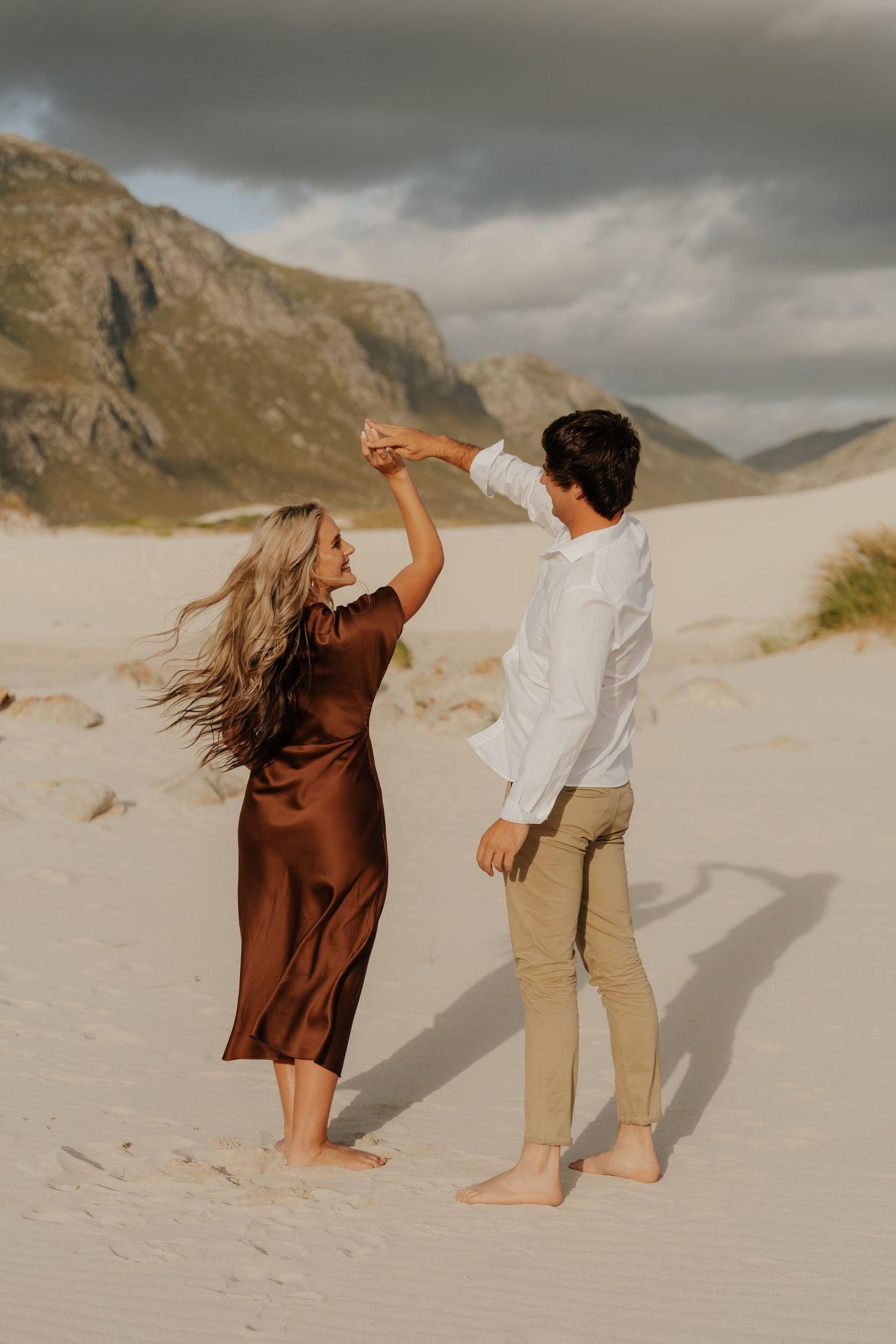 A couple dances barefoot on a sandy beach, woman in brown satin dress, man in white shirt, mountains in background.