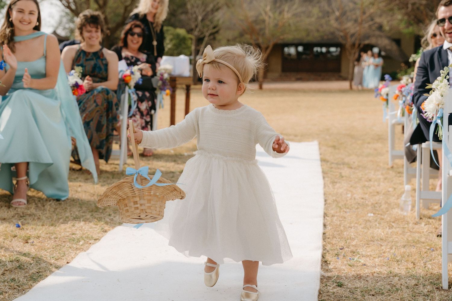 Wedding ceremony guests seated outdoors on white chairs in a rustic setting.
