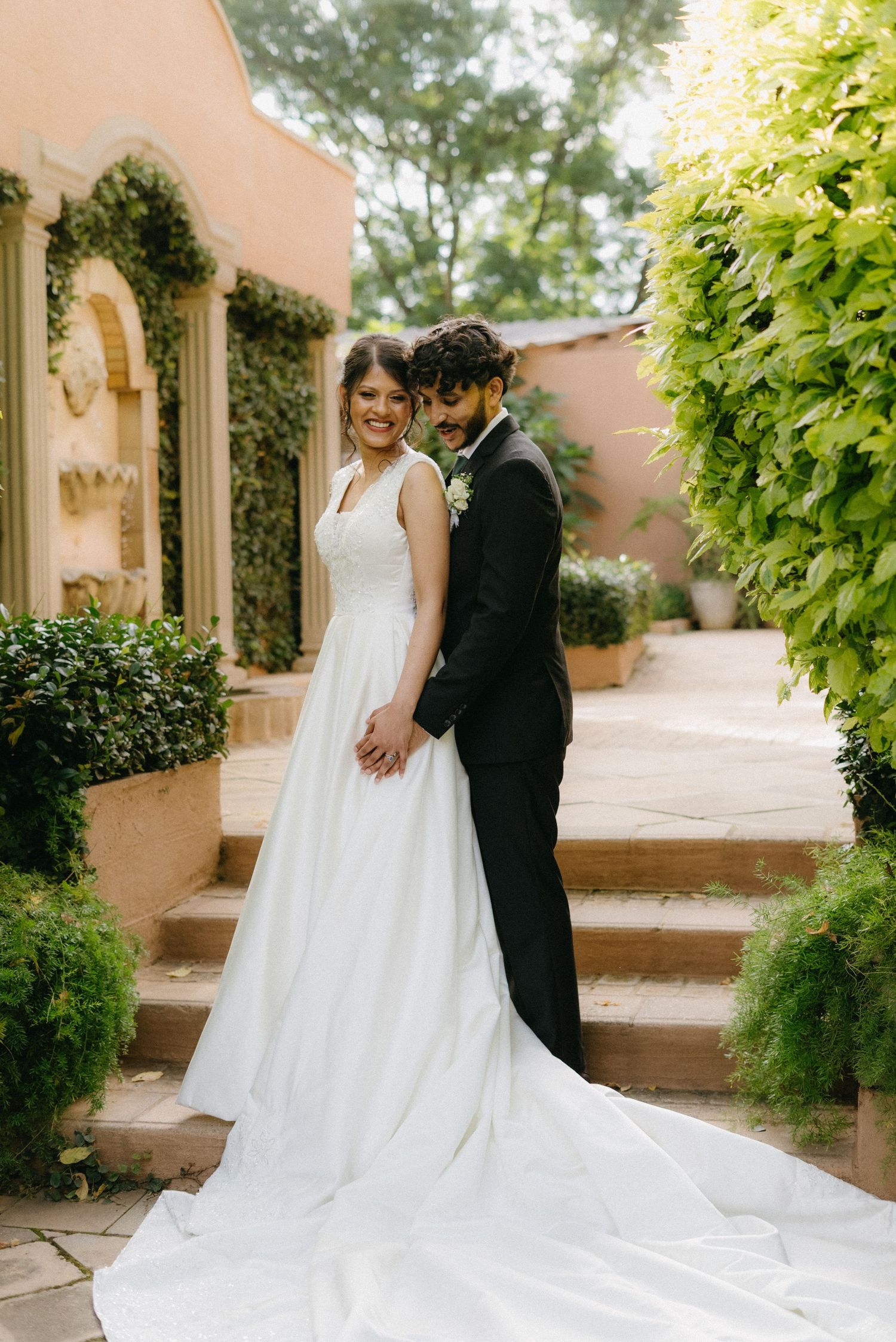 A wedding couple poses along a picturesque garden path with trailing white dress and vine-covered walls.