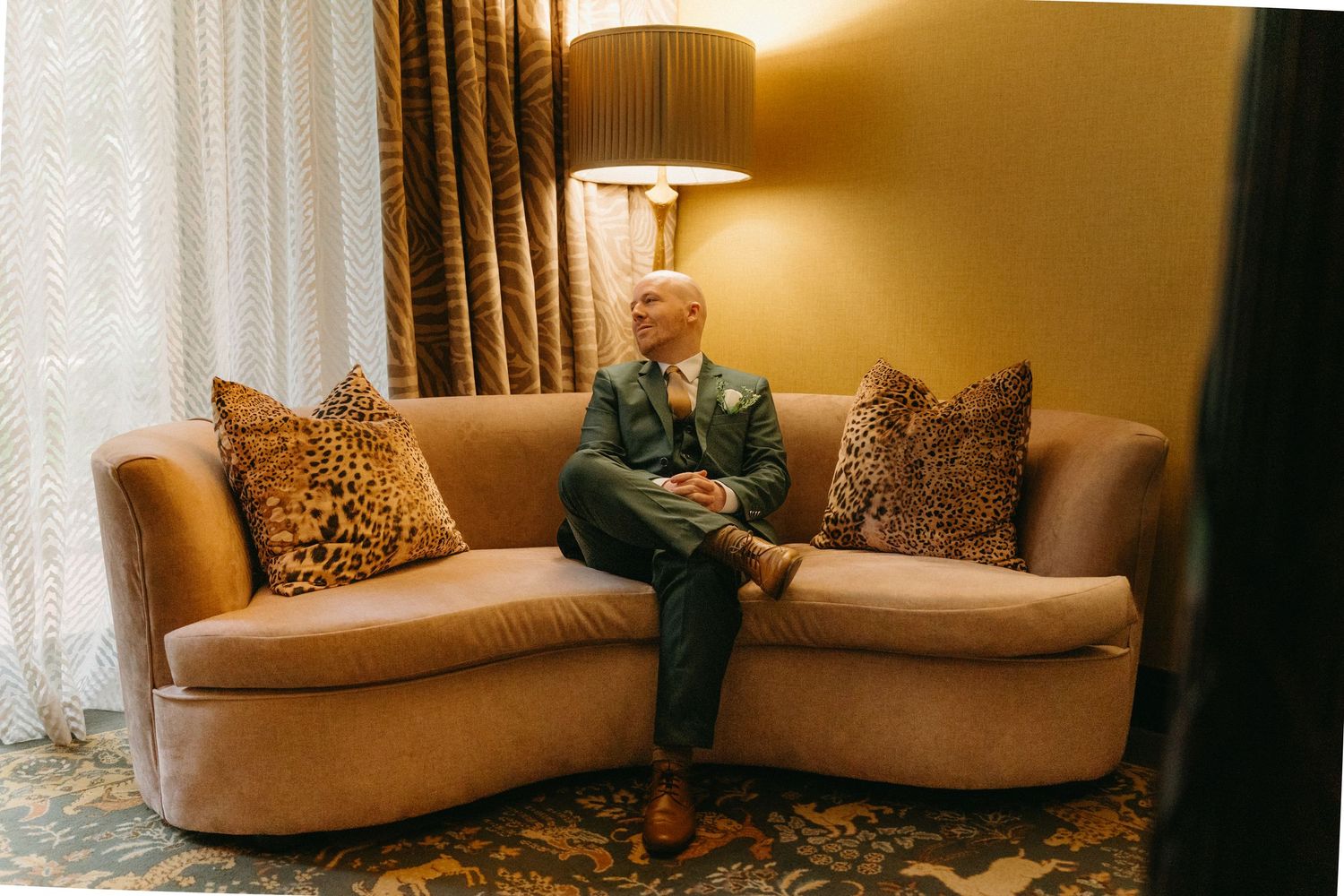 Someone wearing a green suit relaxes on a curved tan couch with leopard print pillows under warm lighting.