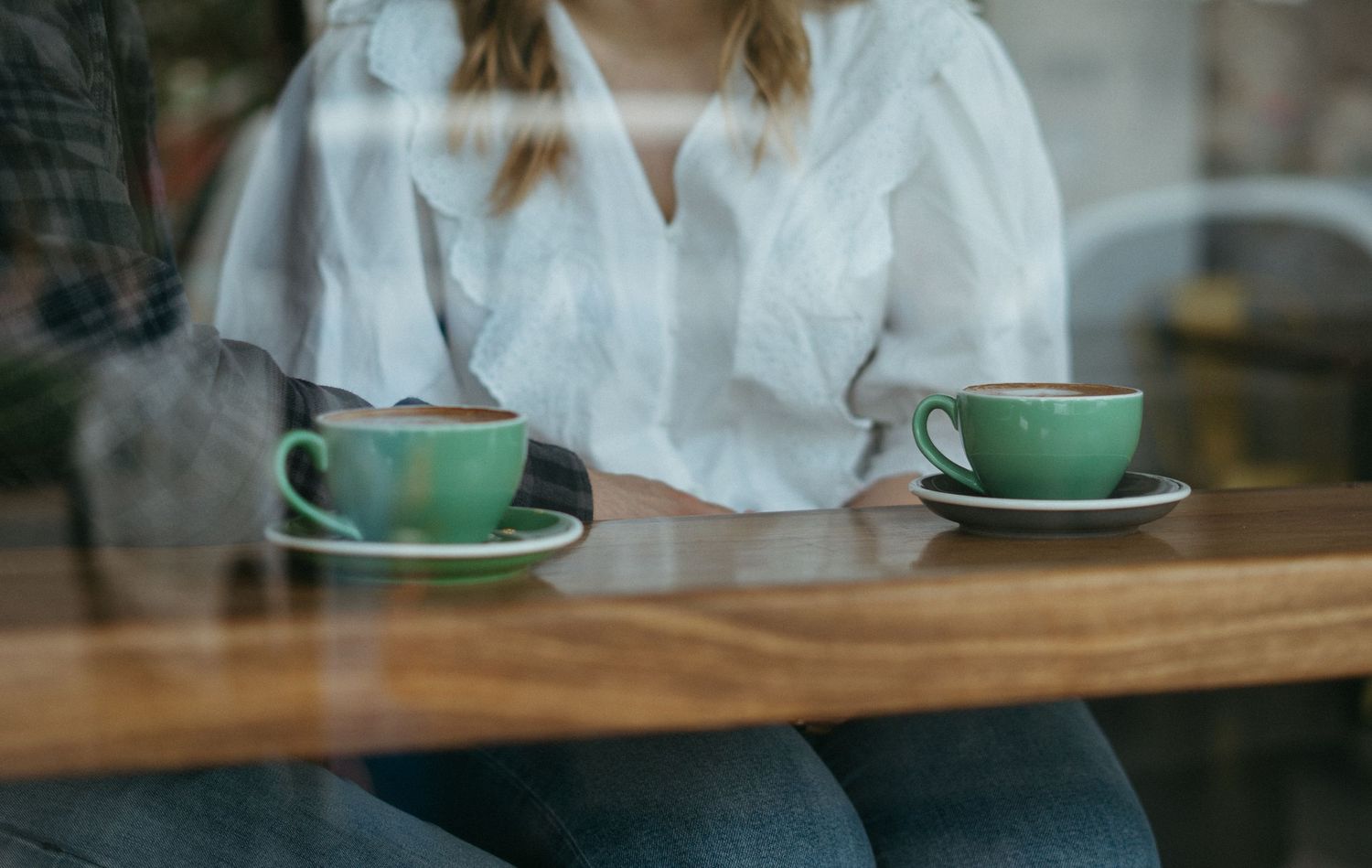 Two green ceramic coffee cups with saucers sit on wooden table with person in white shirt visible through window glass.