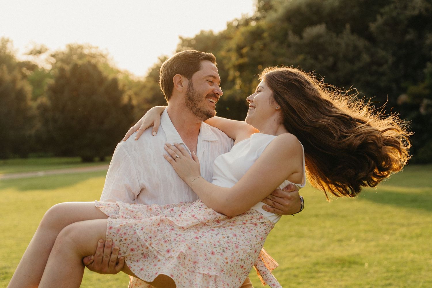 A joyful couple shares a romantic moment outdoors at sunset, man carrying woman in floral skirt on lush green lawn.