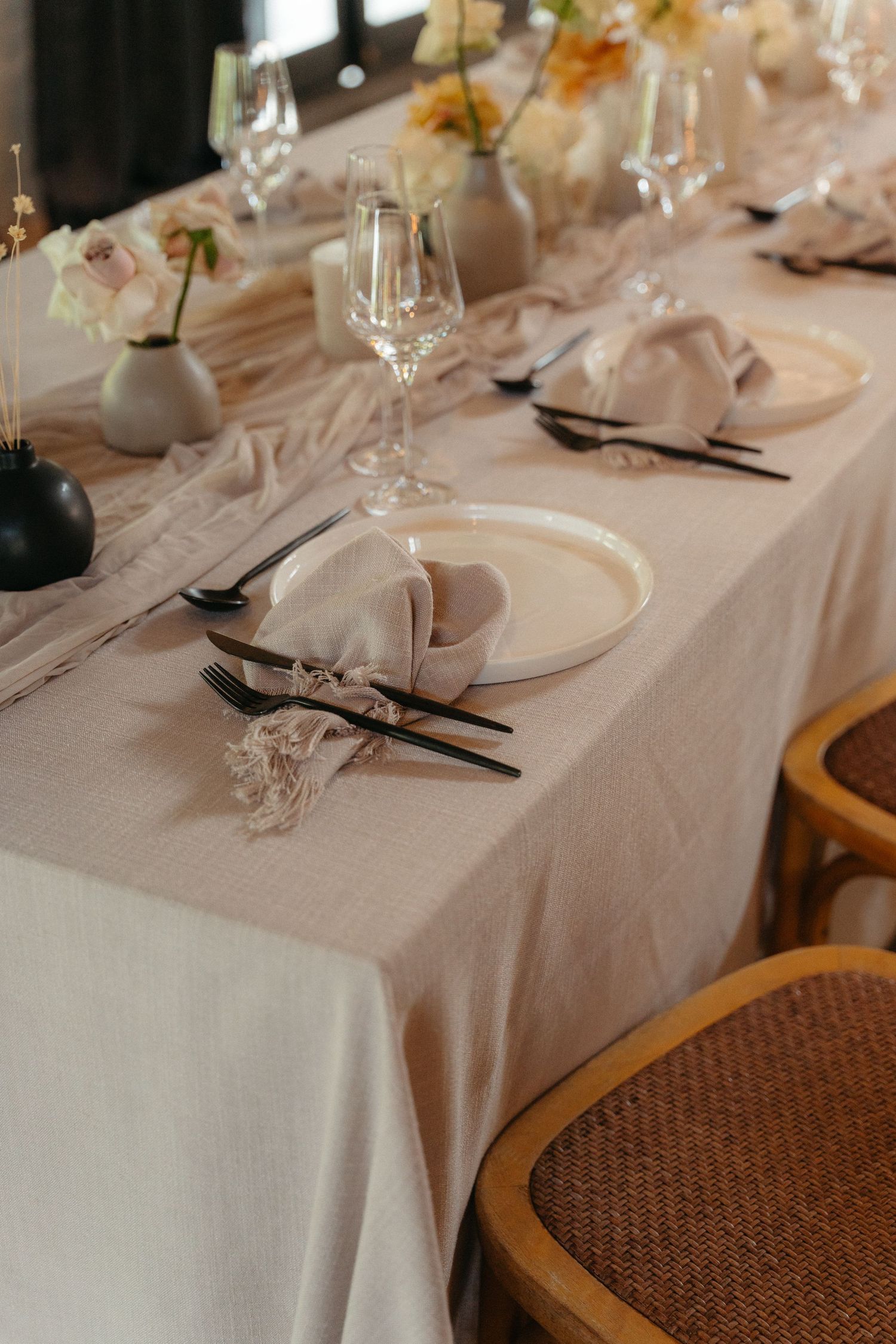 Elegant table setting with white linens, wine glasses, and dark cutlery arranged on a rattan-backed chair.