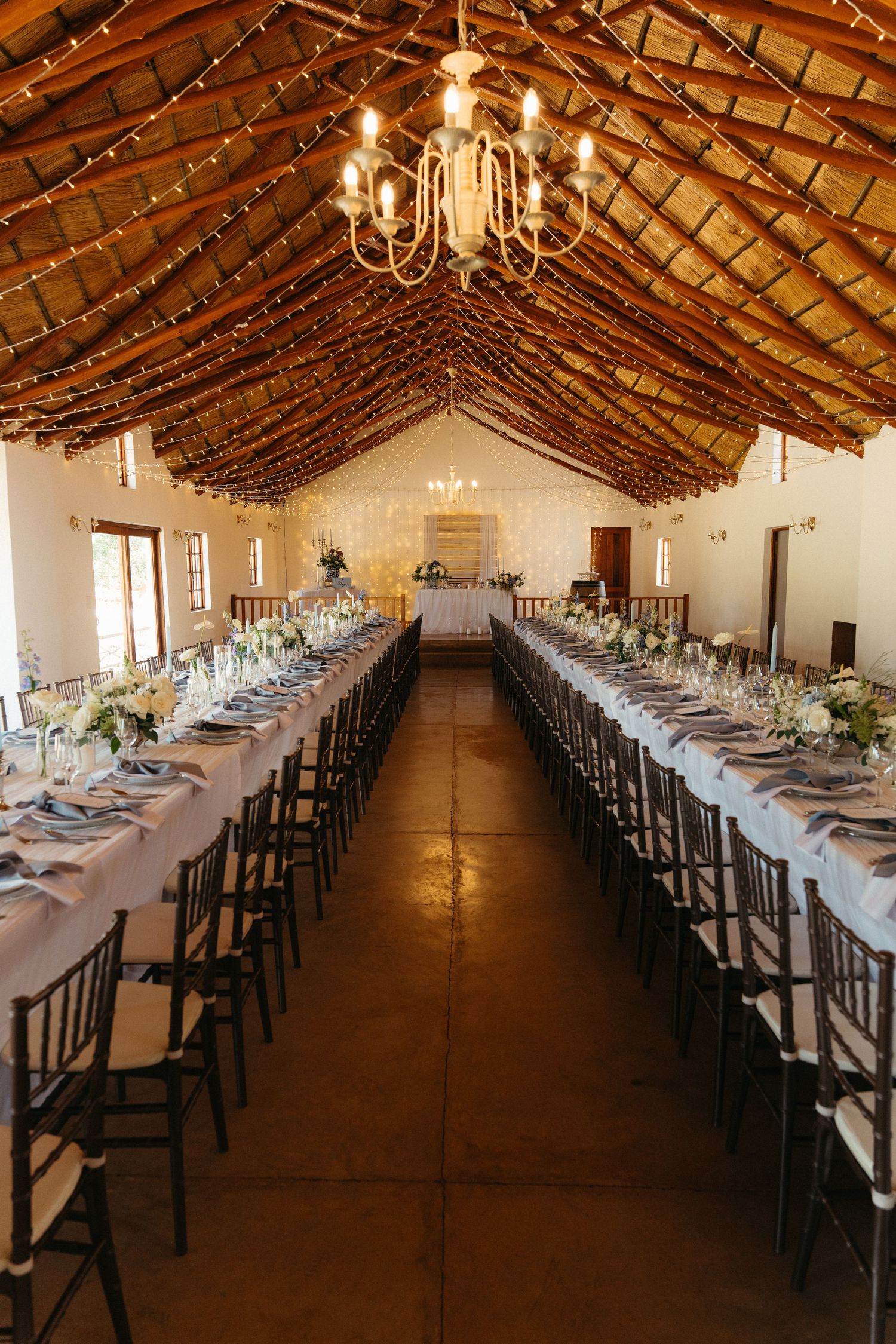 Long rustic reception hall with exposed wooden beams and chandeliers set for elegant dining with white tablecloths.