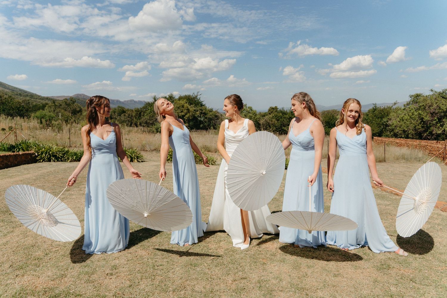 Bridesmaids in light blue dresses twirl and dance together in a scenic outdoor field with mountains in the background.