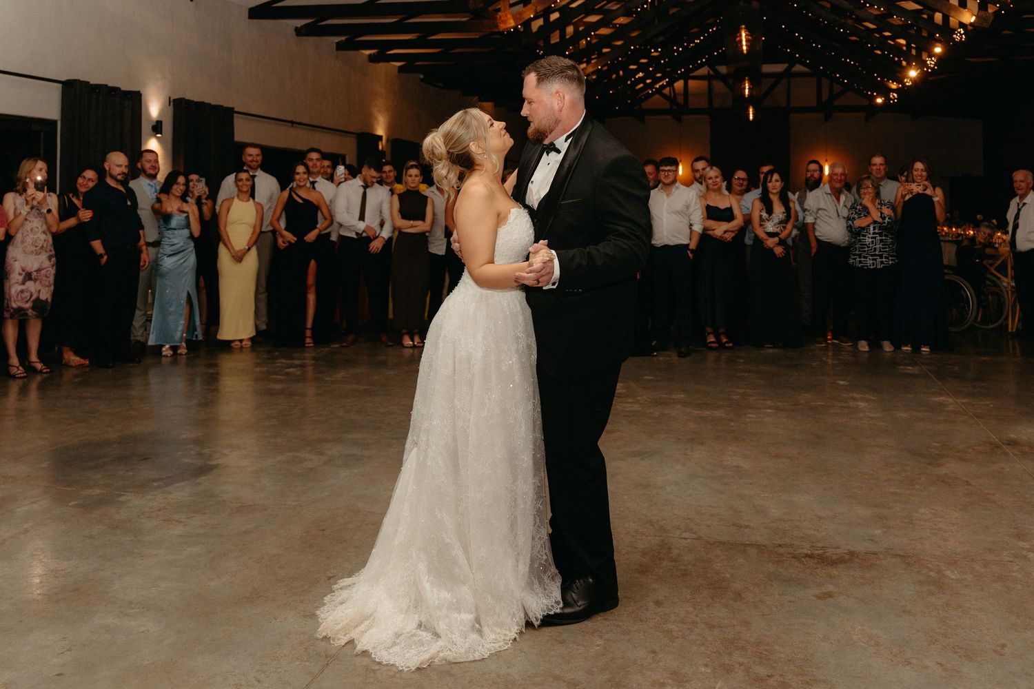 A couple shares their first dance in a dimly lit reception venue with guests watching from the background.