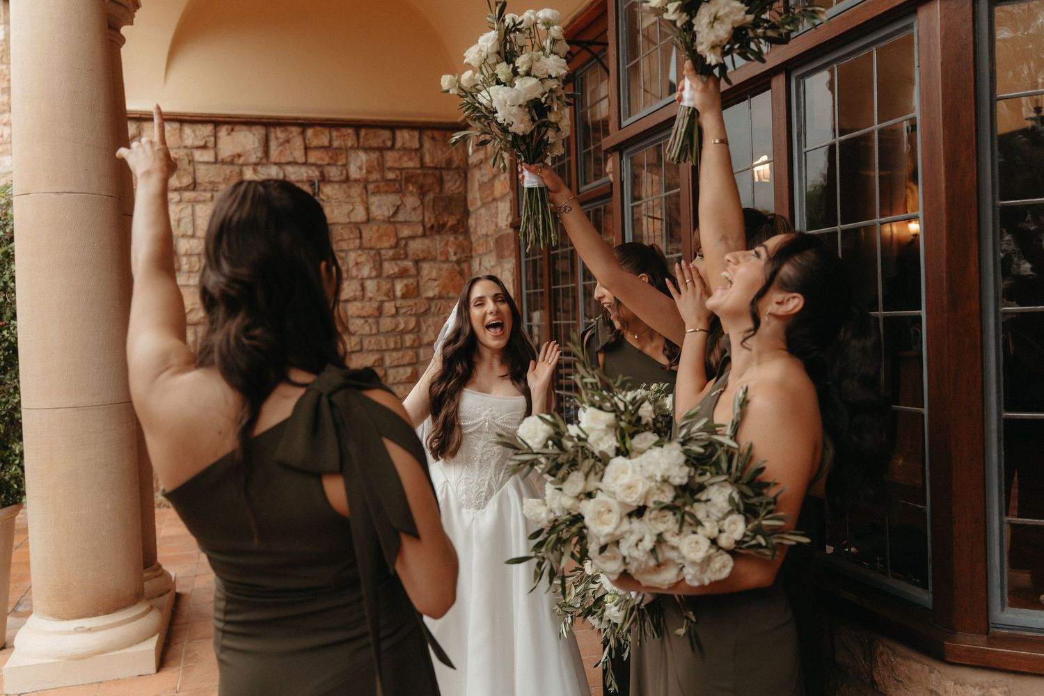 Bride laughing with bridesmaids in olive gowns raising white floral bouquets outside Glenshiel in Johannesburg